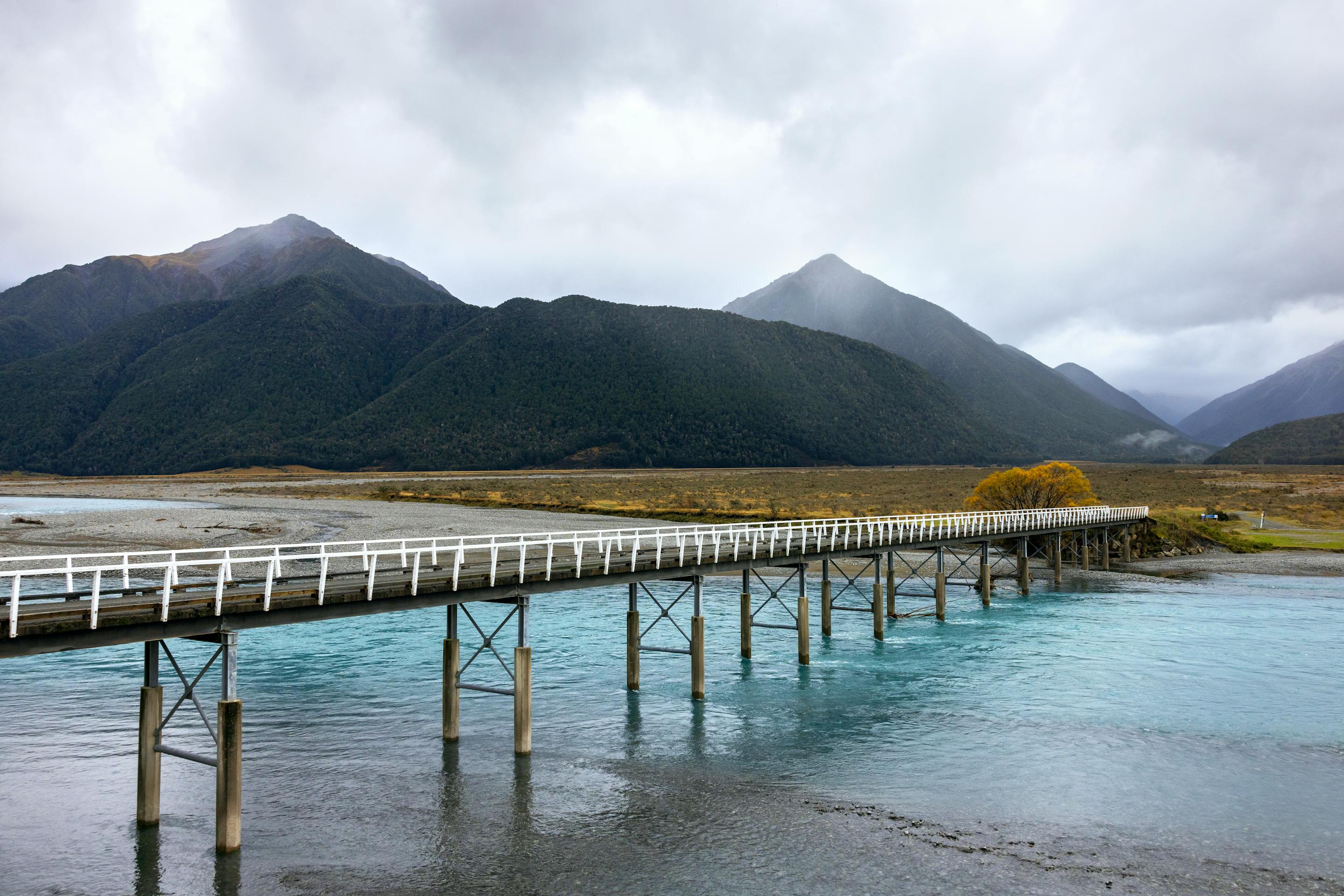 Long wooden pier extends over clear lake water toward dark mountains, with golden grasses and misty light beyond.
