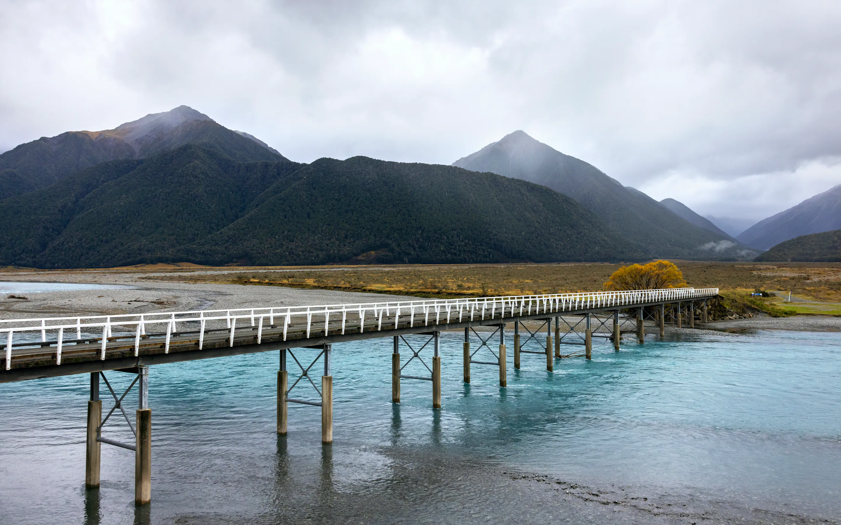 Long wooden pier extends over clear lake water toward dark mountains, with golden grasses and misty light beyond.