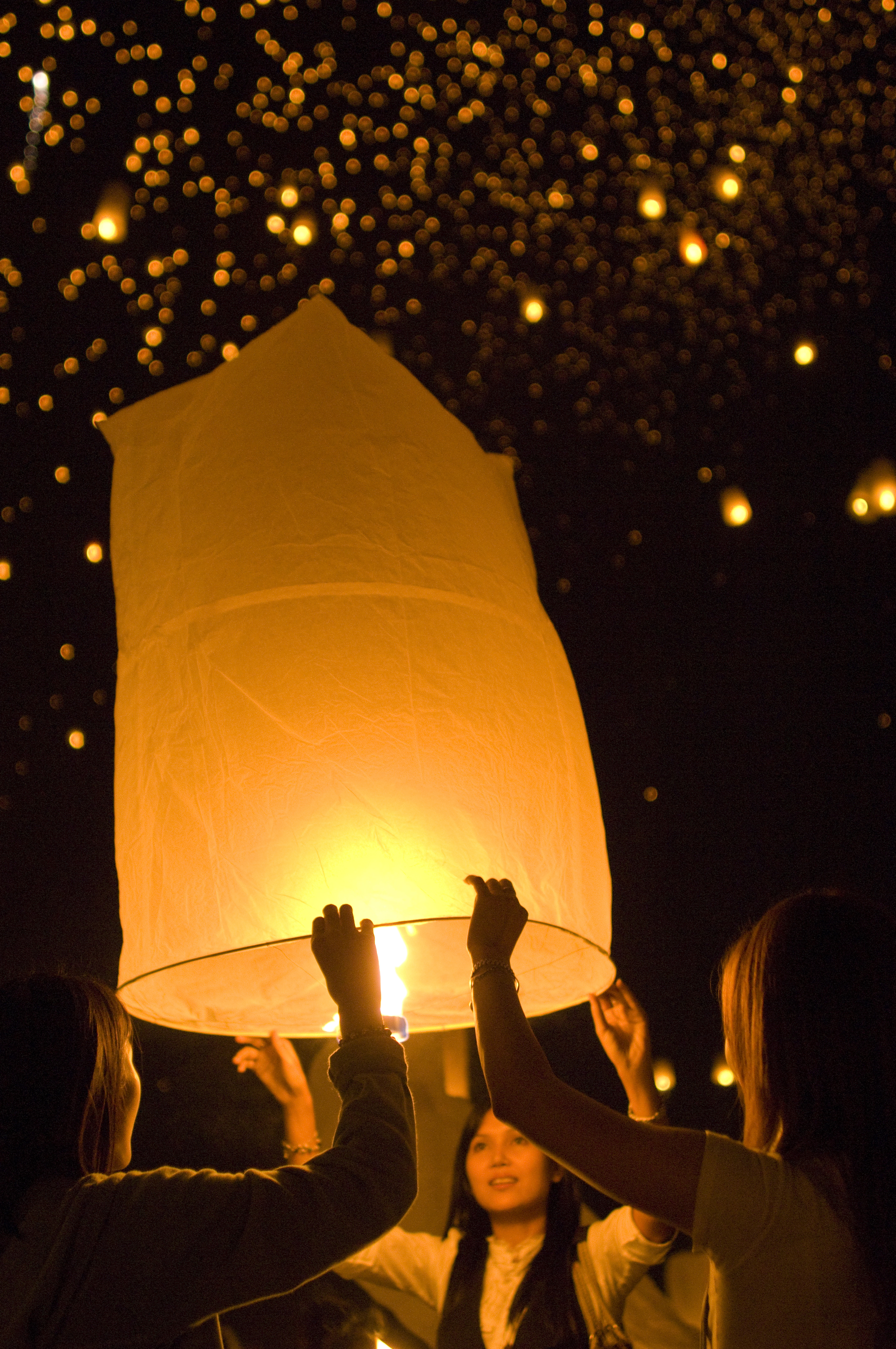 A glowing paper sky lantern rises at night as people lift it overhead, sparks and bokeh lights behind softly.