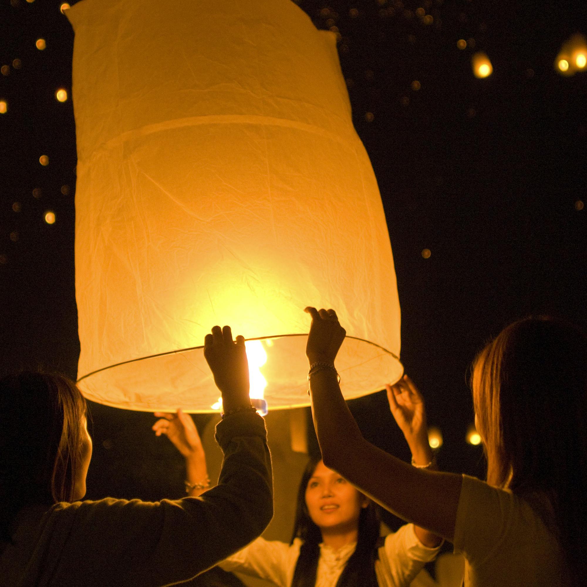 A glowing paper sky lantern rises at night as people lift it overhead, sparks and bokeh lights behind softly.