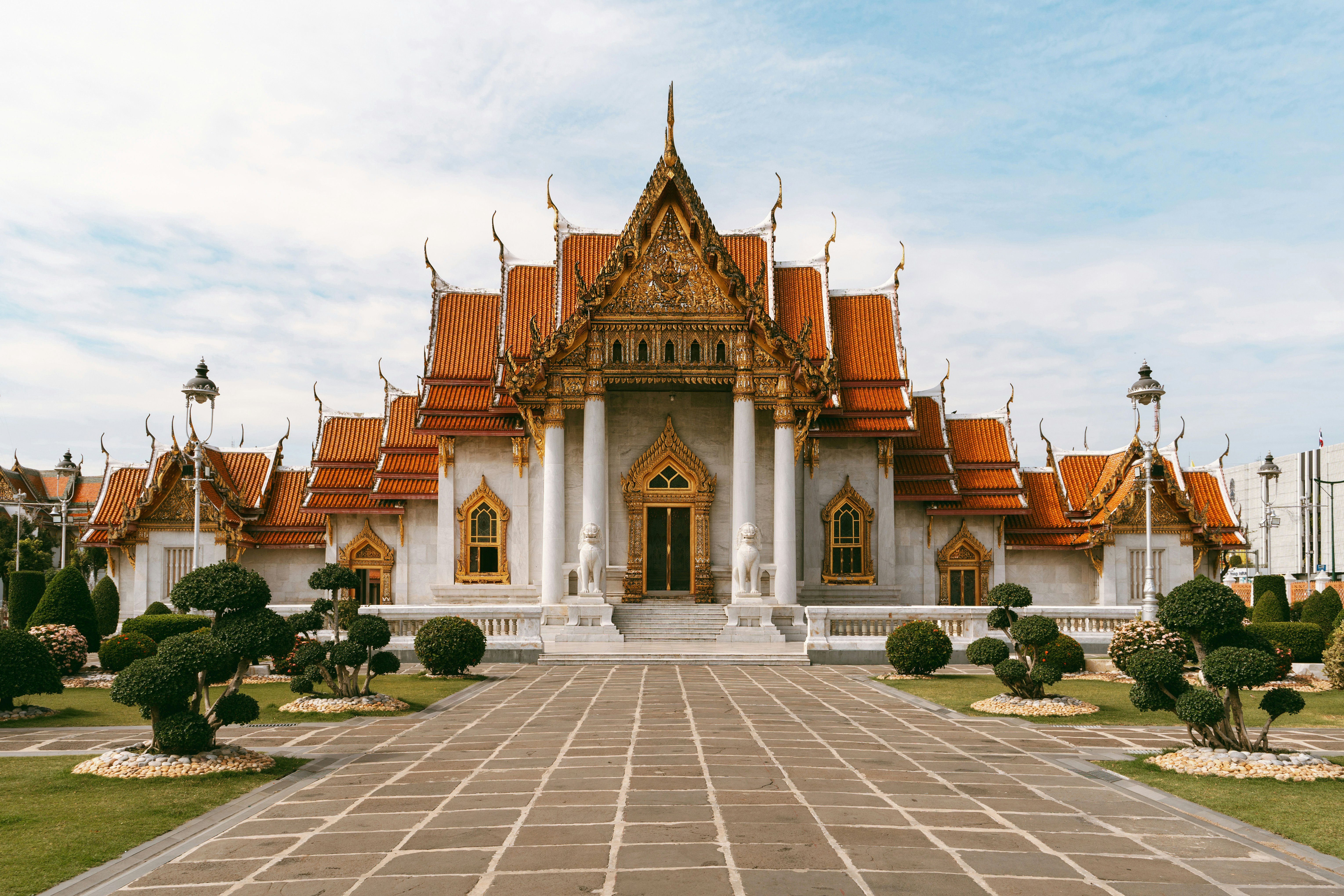 An ornate Thai temple stands at the end of a stone walkway, with manicured shrubs and cloudy sky overhead.