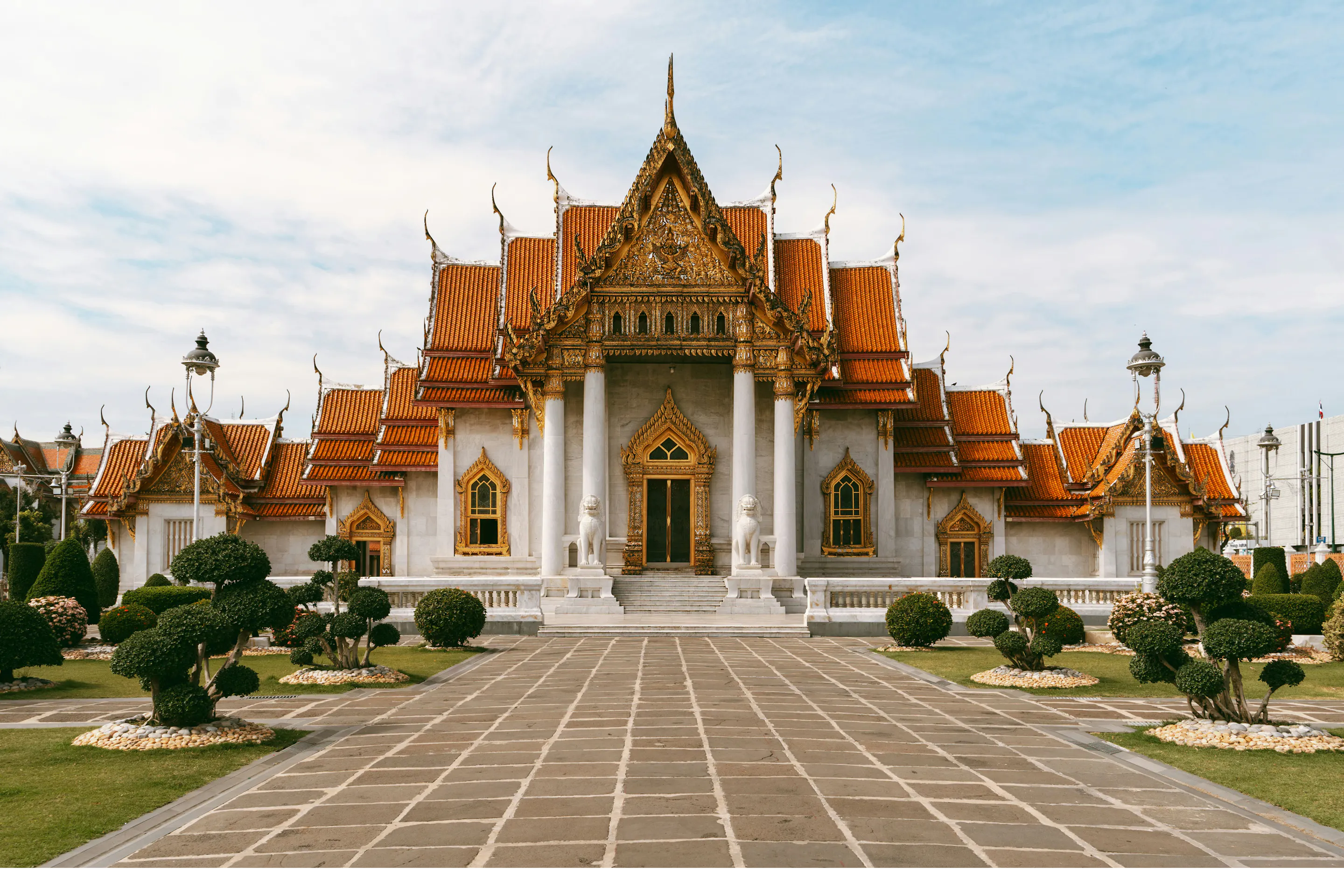 An ornate Thai temple stands at the end of a stone walkway, with manicured shrubs and cloudy sky overhead.