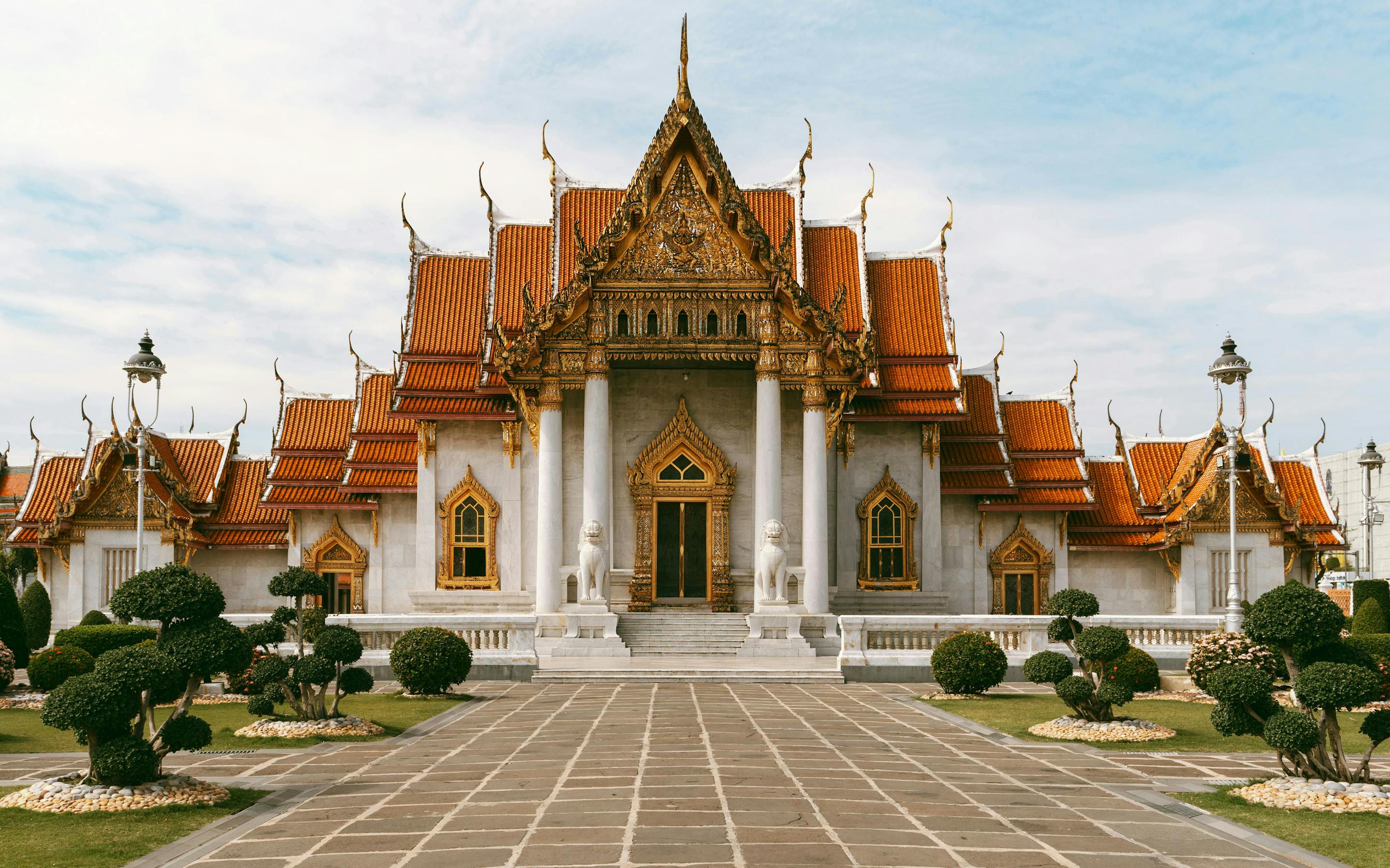 An ornate Thai temple stands at the end of a stone walkway, with manicured shrubs and cloudy sky overhead.