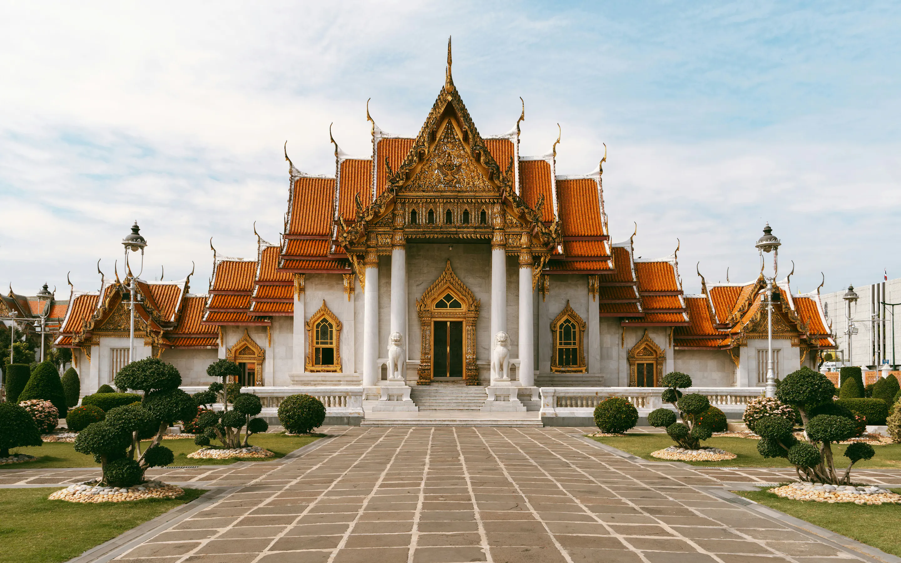 An ornate Thai temple stands at the end of a stone walkway, with manicured shrubs and cloudy sky overhead.