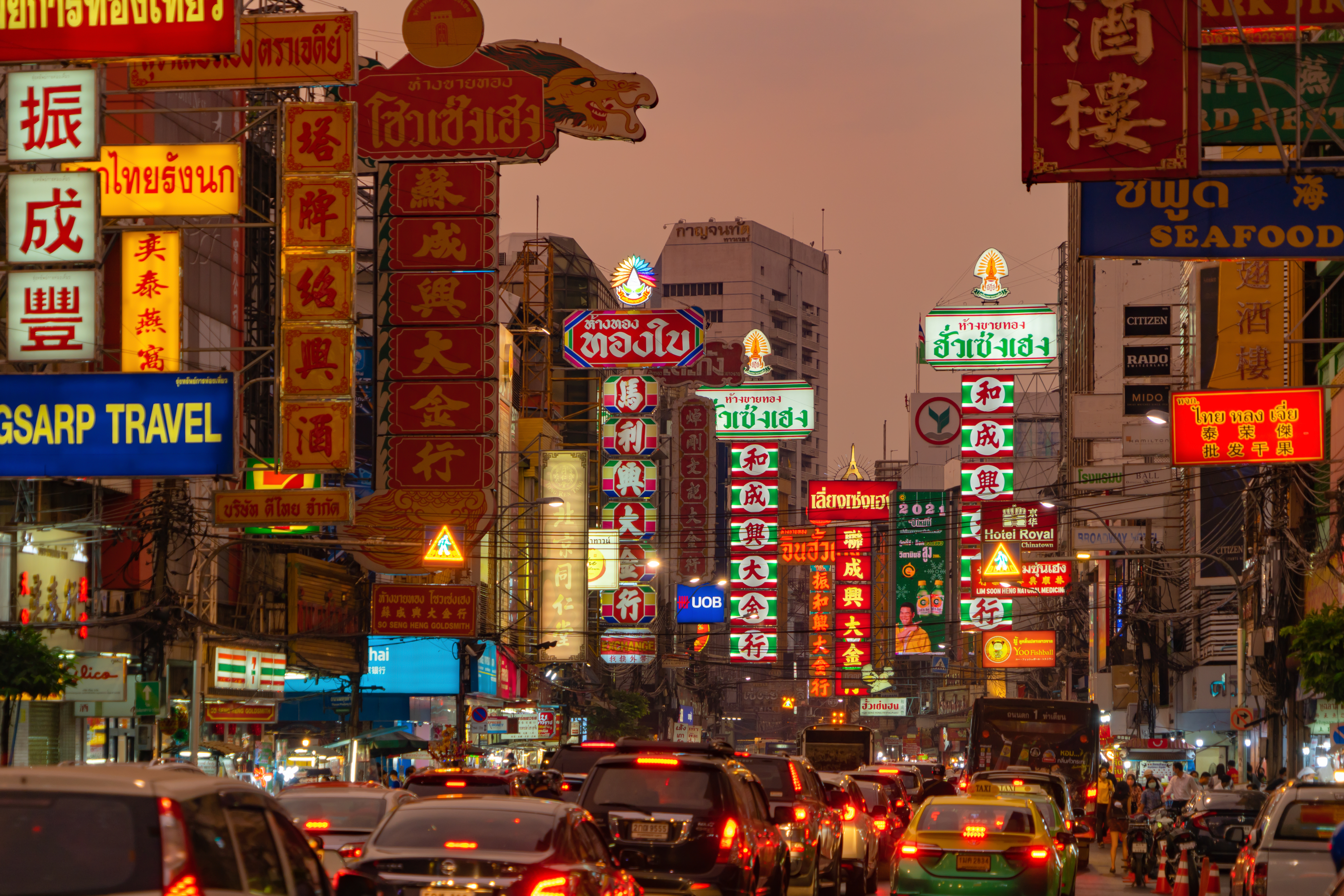 Traffic crawls through Bangkok's Chinatown at dusk, neon signs glowing above the crowded street and taxis.