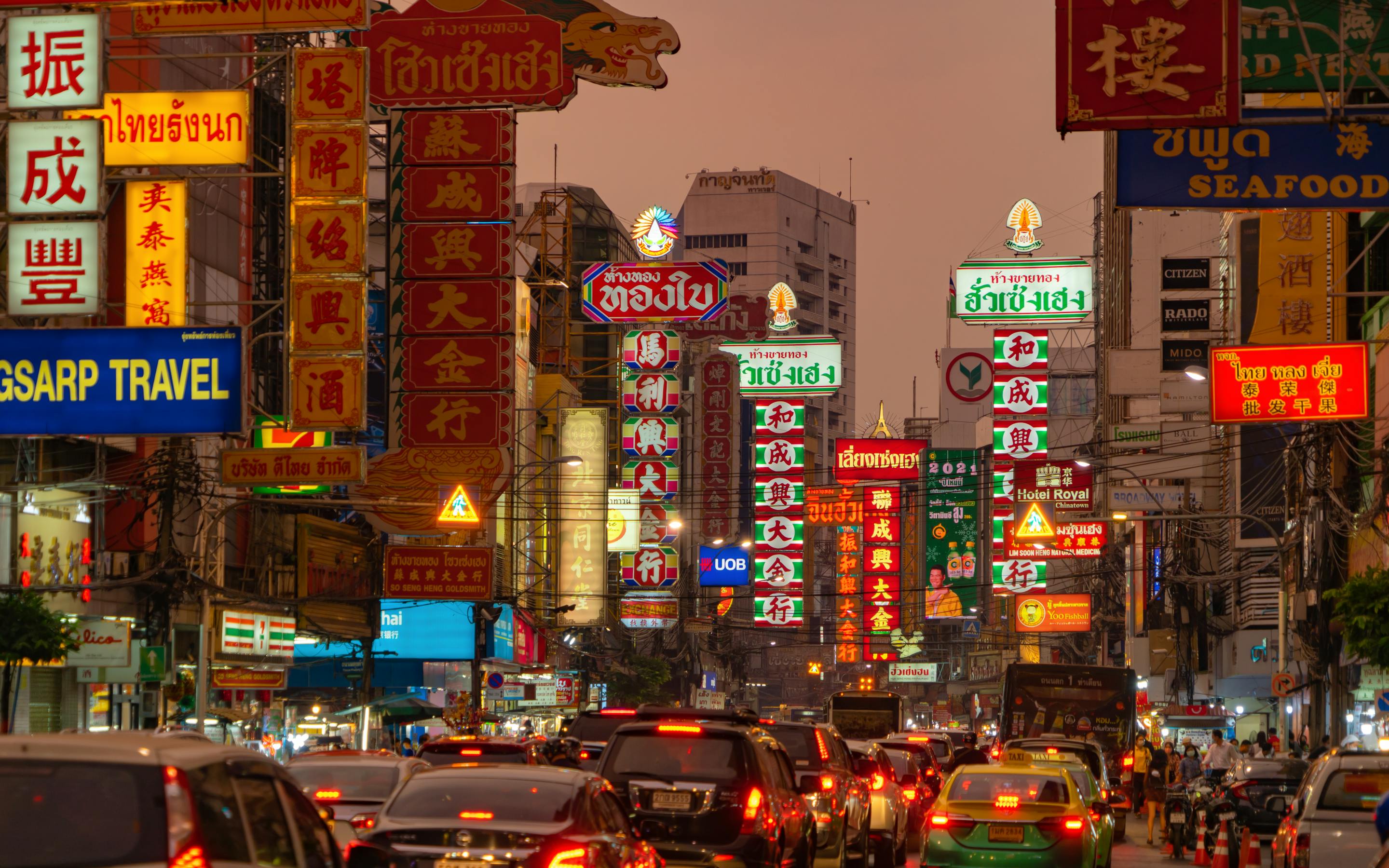 Traffic crawls through Bangkok's Chinatown at dusk, neon signs glowing above the crowded street and taxis.
