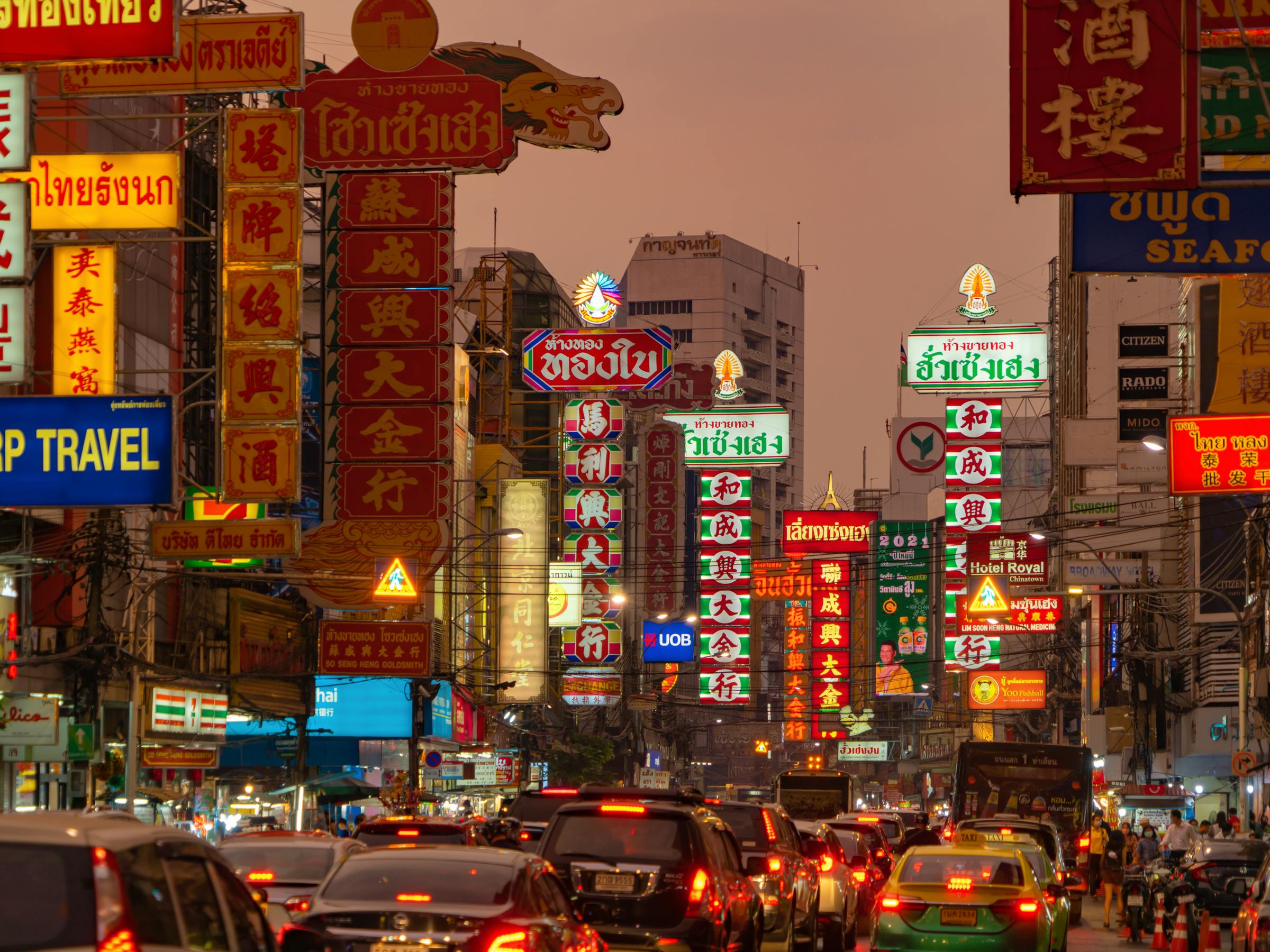 Traffic crawls through Bangkok's Chinatown at dusk, neon signs glowing above the crowded street and taxis.