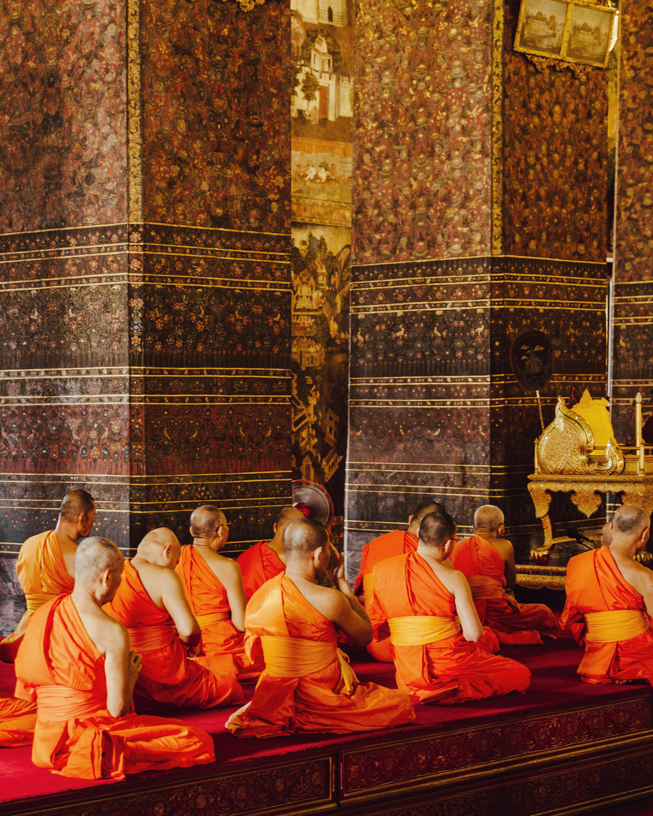 Monks in orange robes sit along an ornate temple wall, framed by patterned tiles and gilded statues inside.