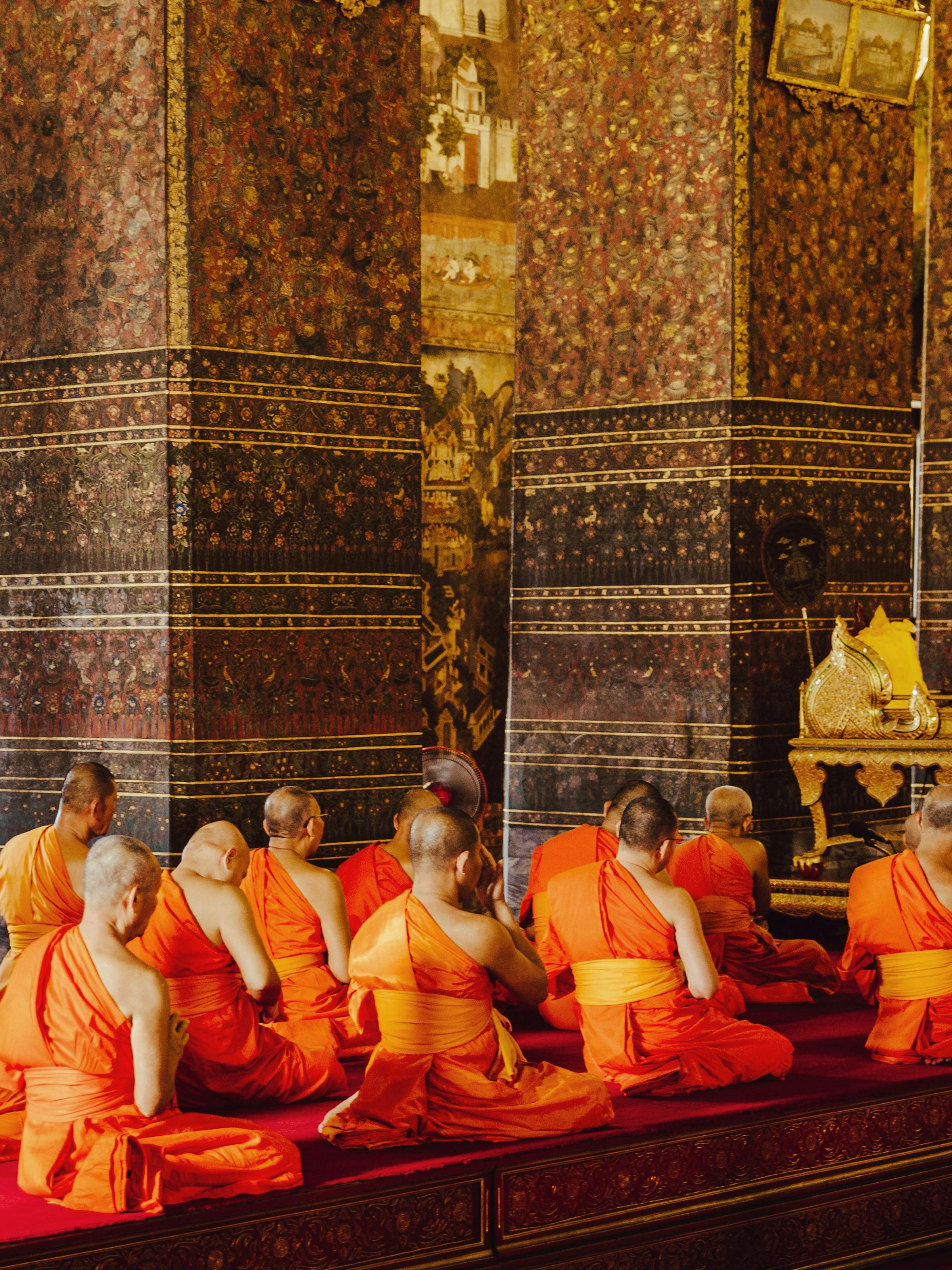 Monks in orange robes sit along an ornate temple wall, framed by patterned tiles and gilded statues inside.