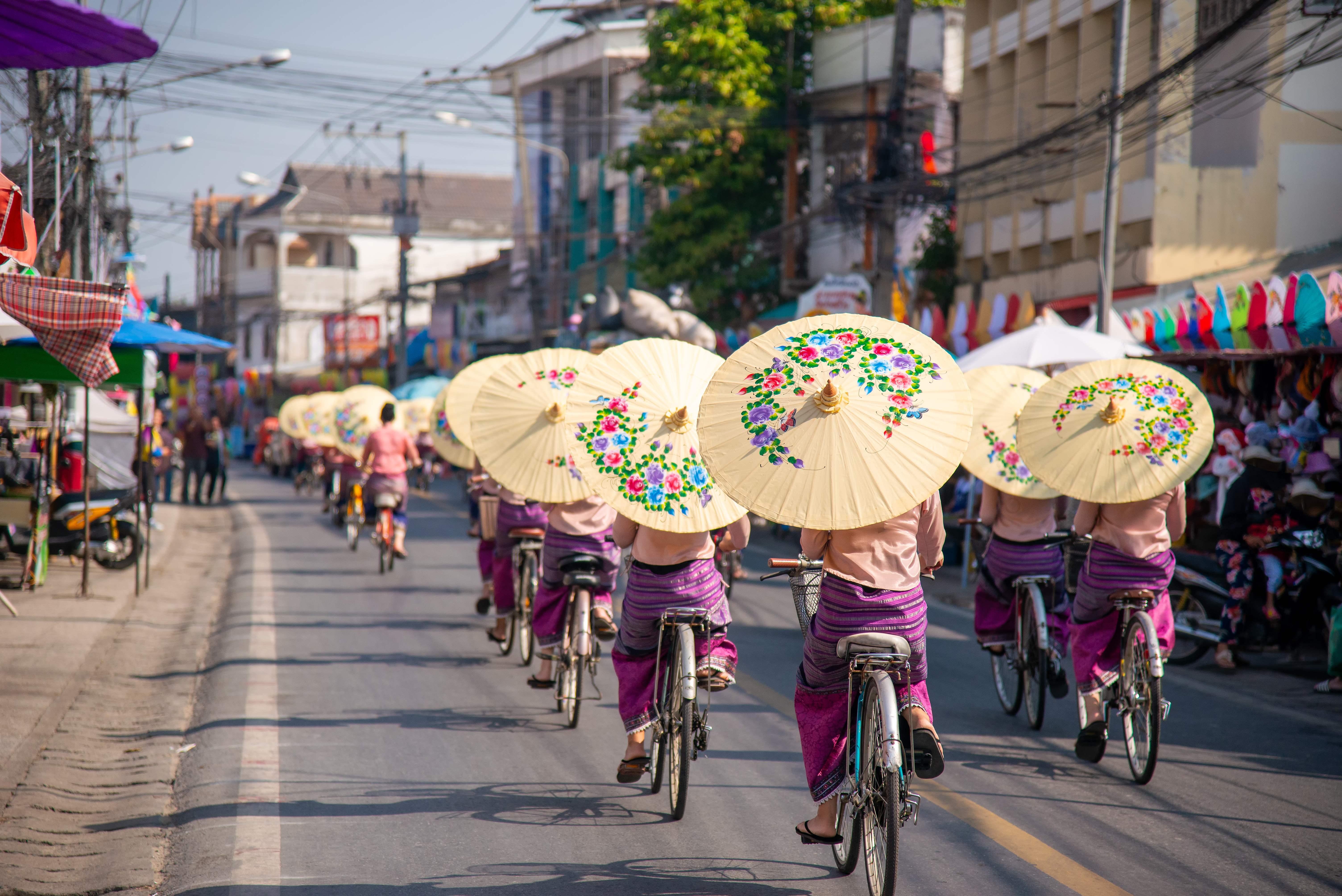 Cyclists ride along a market street carrying painted paper umbrellas, with shops and power lines in the background.