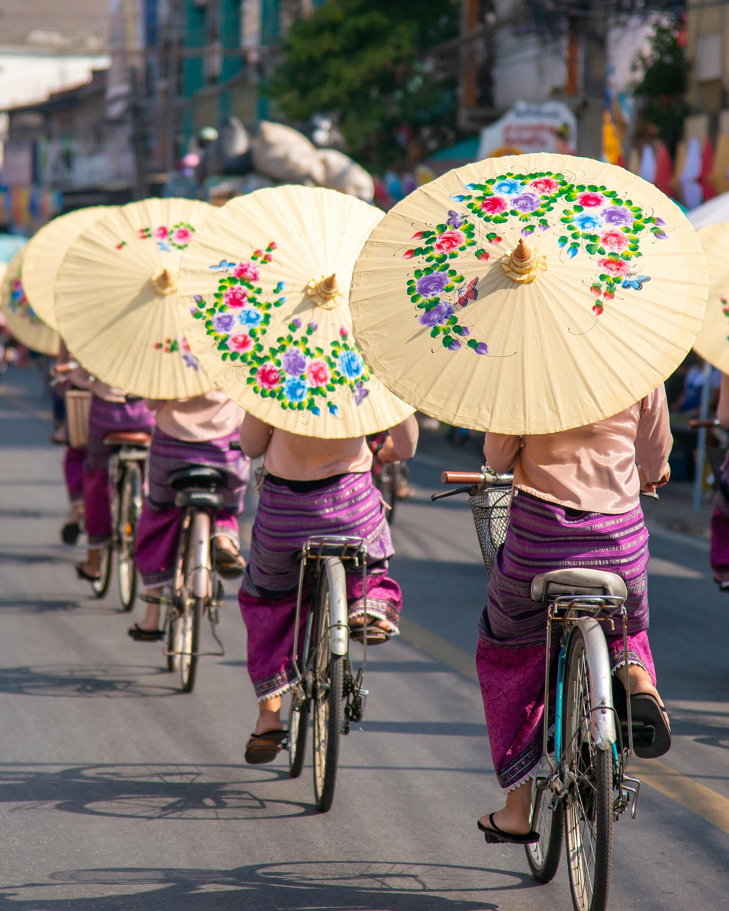 Cyclists ride along a market street carrying painted paper umbrellas, with shops and power lines in the background.