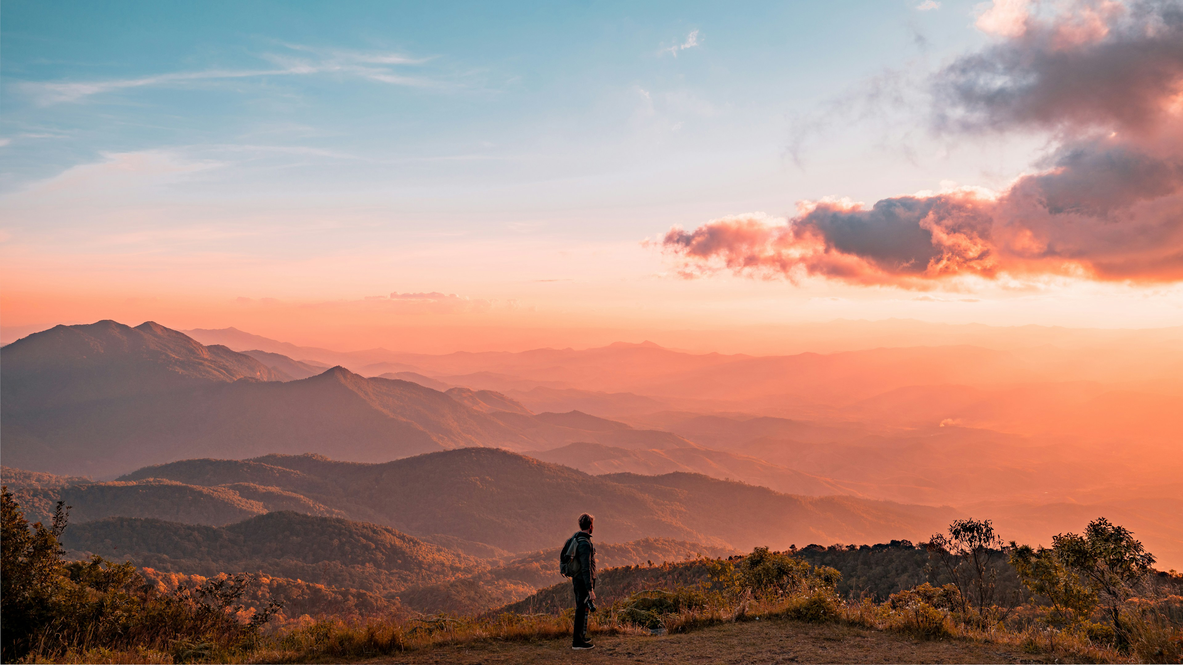 A lone person stands on a ridge at sunset, looking over hazy mountain layers under pink and orange clouds.