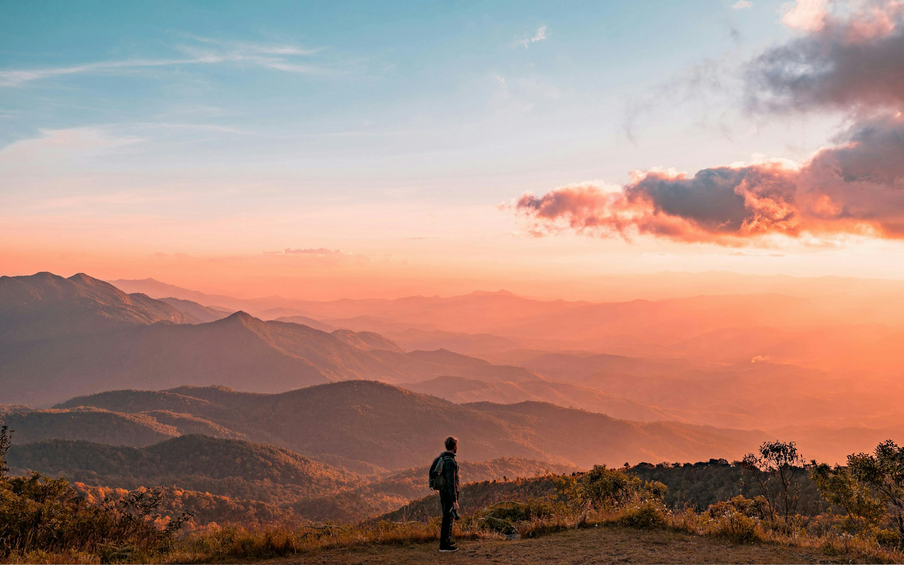 A lone person stands on a ridge at sunset, looking over hazy mountain layers under pink and orange clouds.