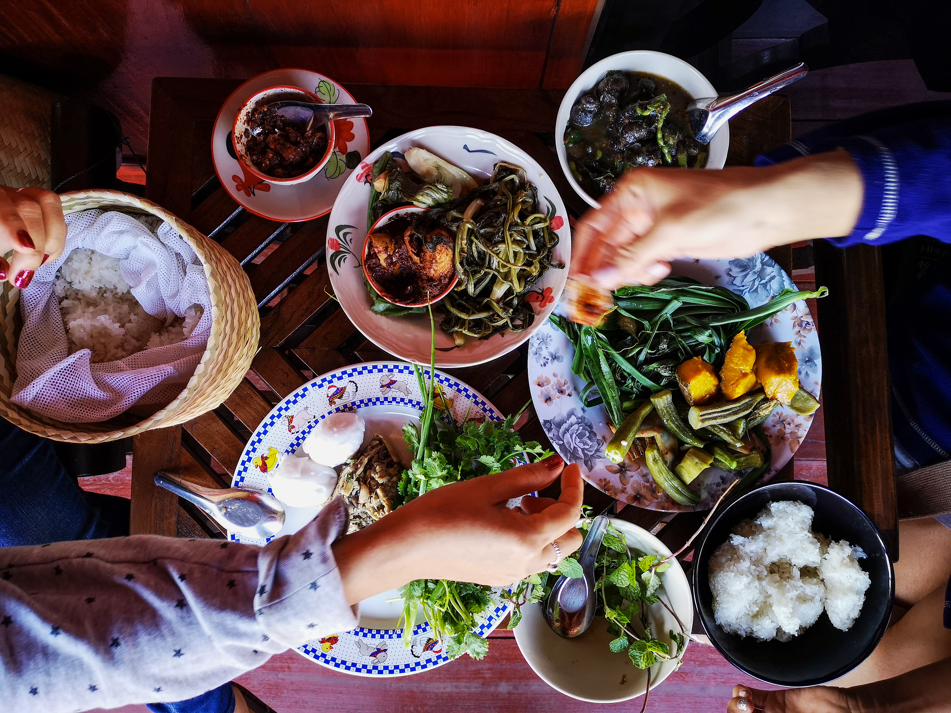 Hands reach over a table of Thai dishes, including curries, vegetables, rice, and bowls set on dark wood.
