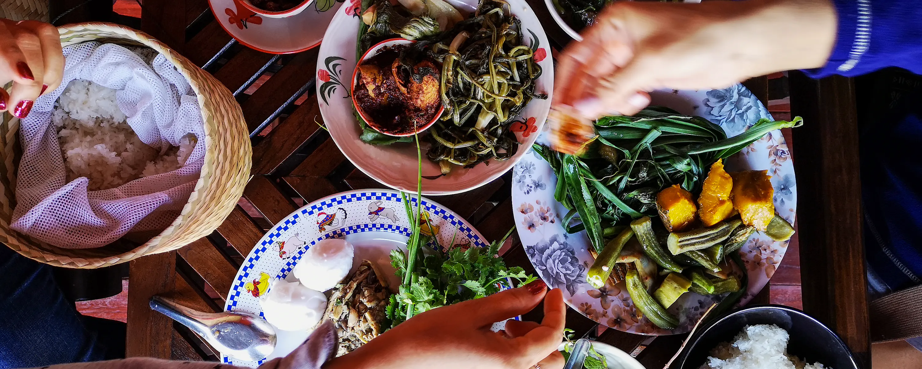 Hands reach over a table of Thai dishes, including curries, vegetables, rice, and bowls set on dark wood.