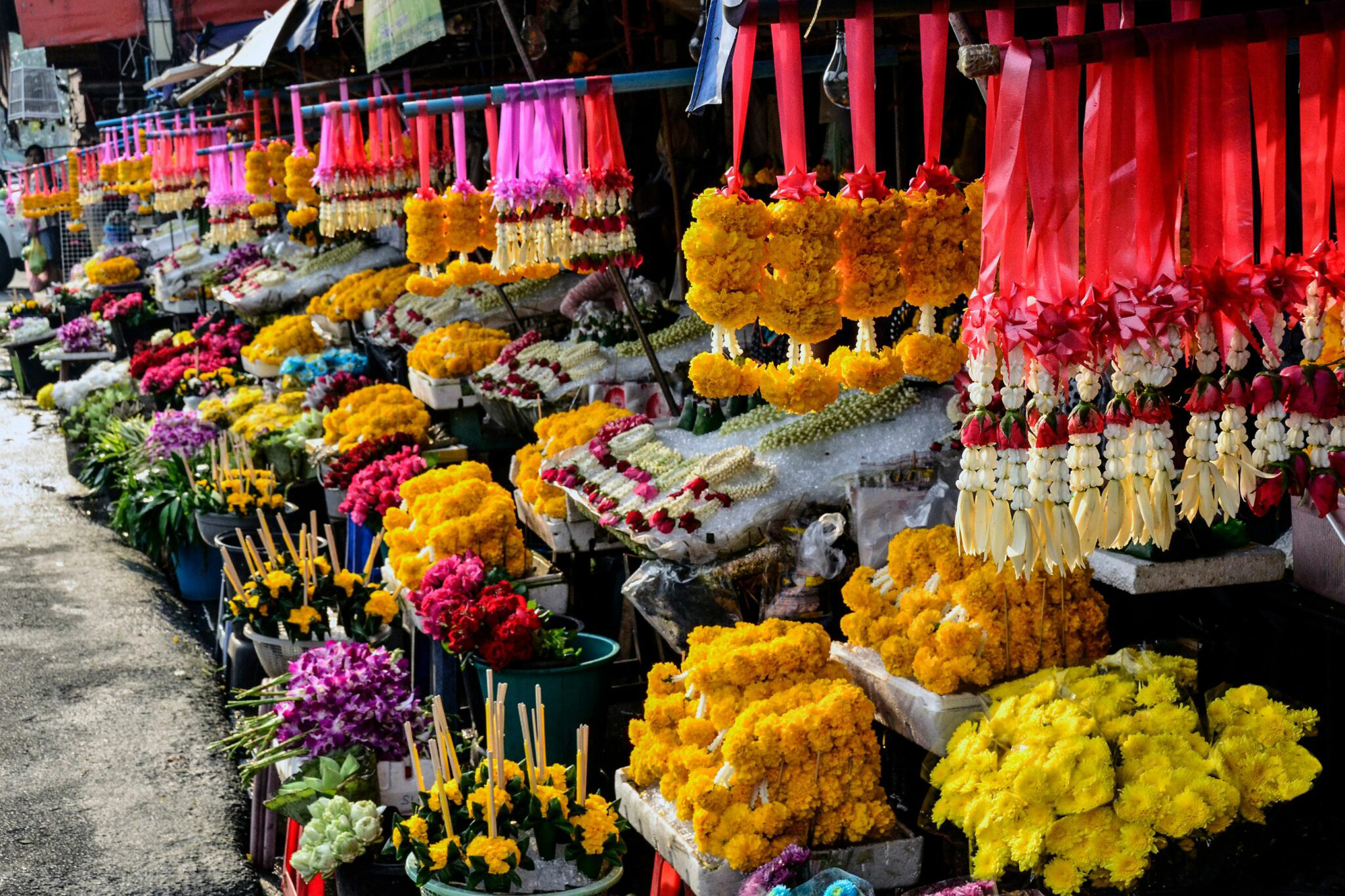 Bright flower market stall with hanging garlands and baskets of colorful blooms lined along a busy walkway.