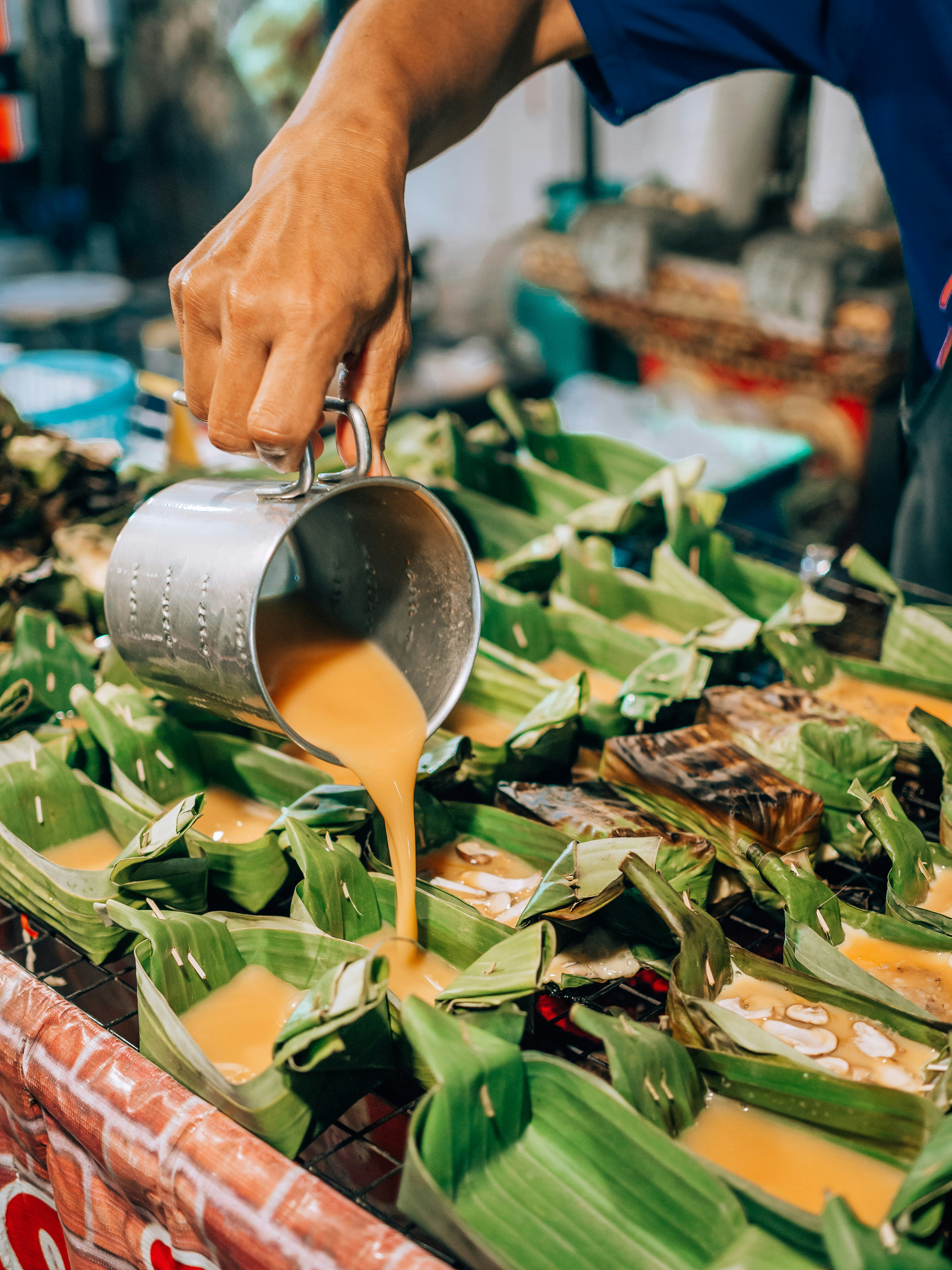 A vendor pours orange curry from a metal pot onto banana-leaf parcels arranged on a busy street food counter.