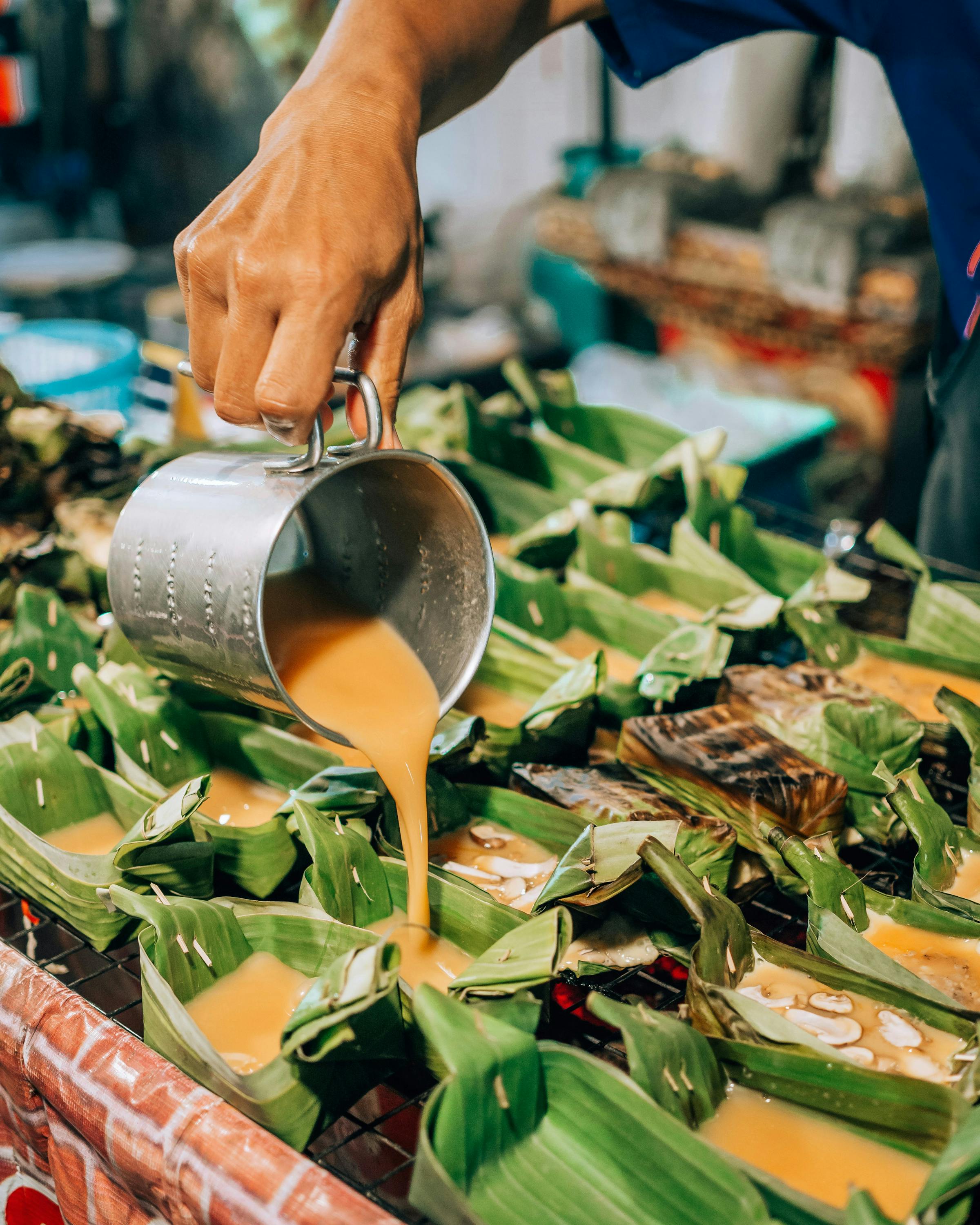 A vendor pours orange curry from a metal pot onto banana-leaf parcels arranged on a busy street food counter.