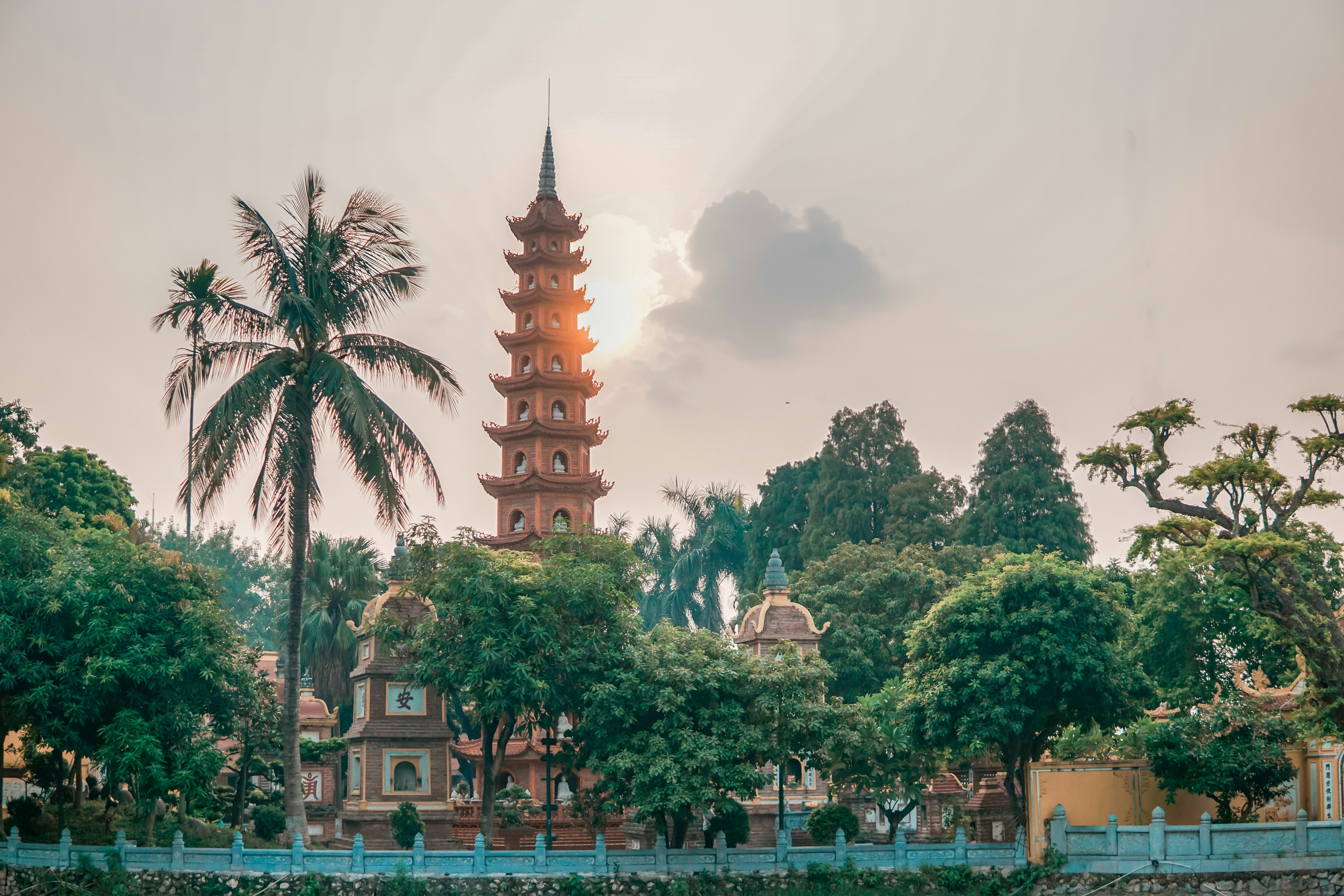 A tall pagoda tower rises above trees at quiet dusk, with a palm frond and hazy sky framing the Hanoi skyline.