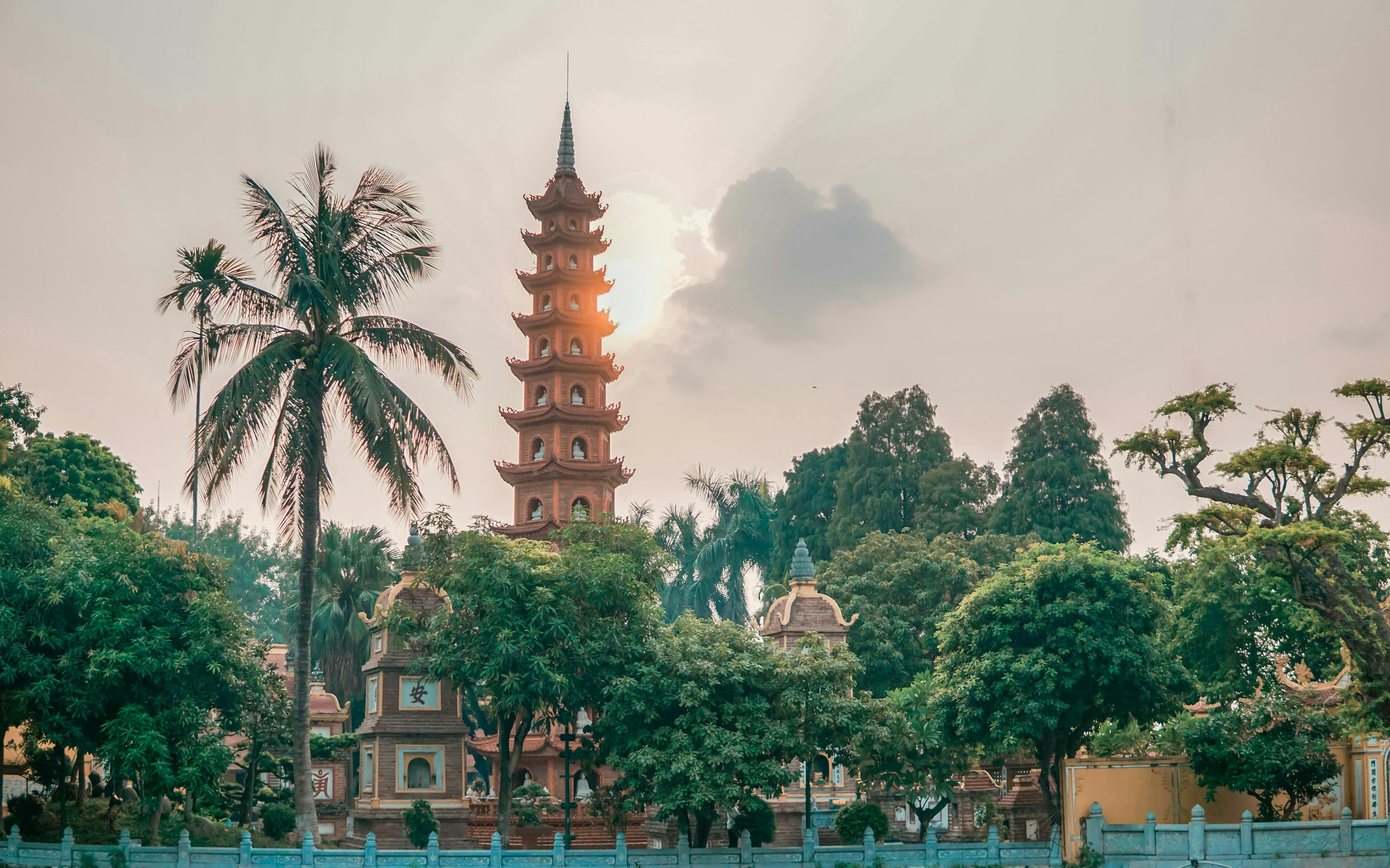 A tall pagoda tower rises above trees at quiet dusk, with a palm frond and hazy sky framing the Hanoi skyline.