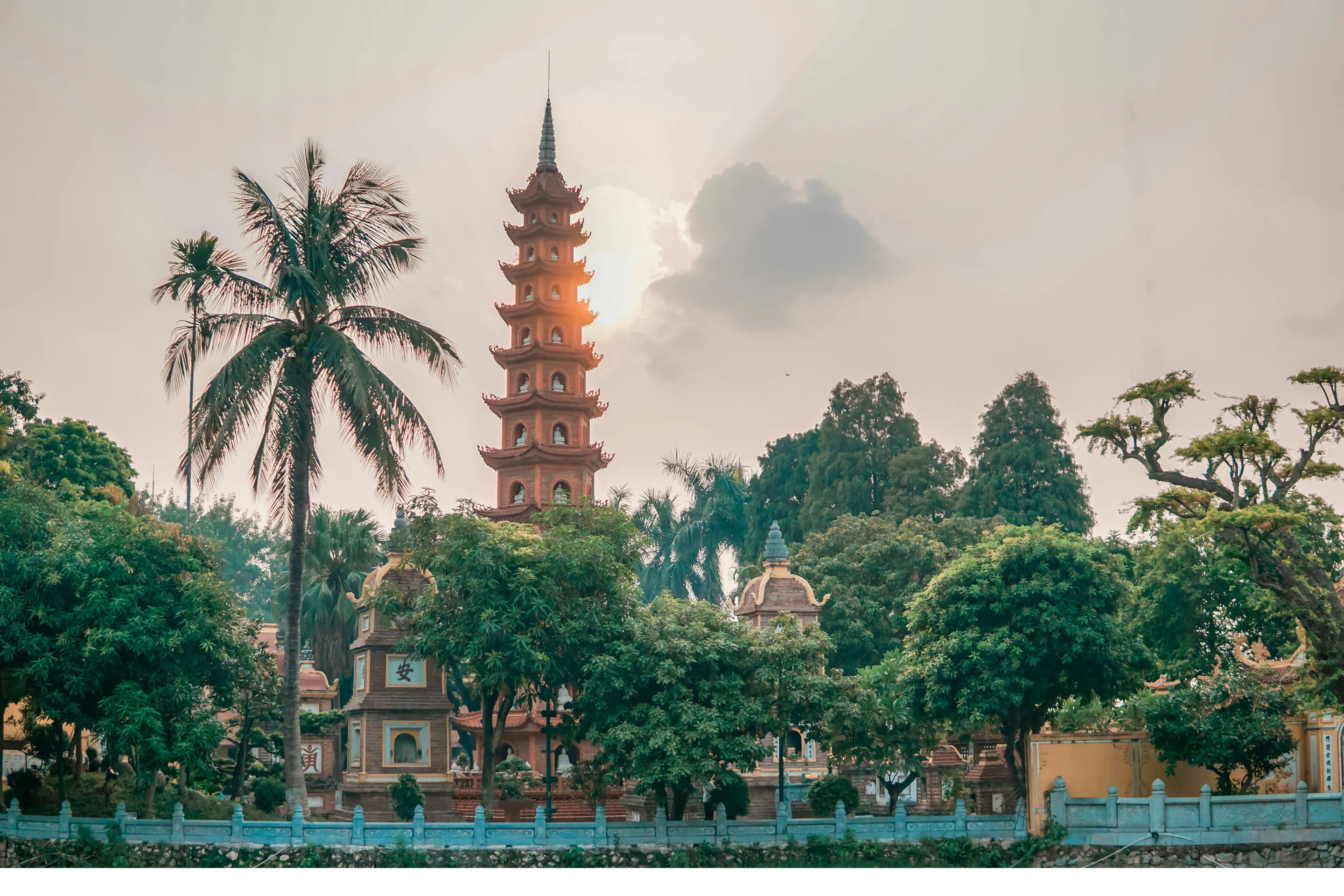 A tall pagoda tower rises above trees at quiet dusk, with a palm frond and hazy sky framing the Hanoi skyline.