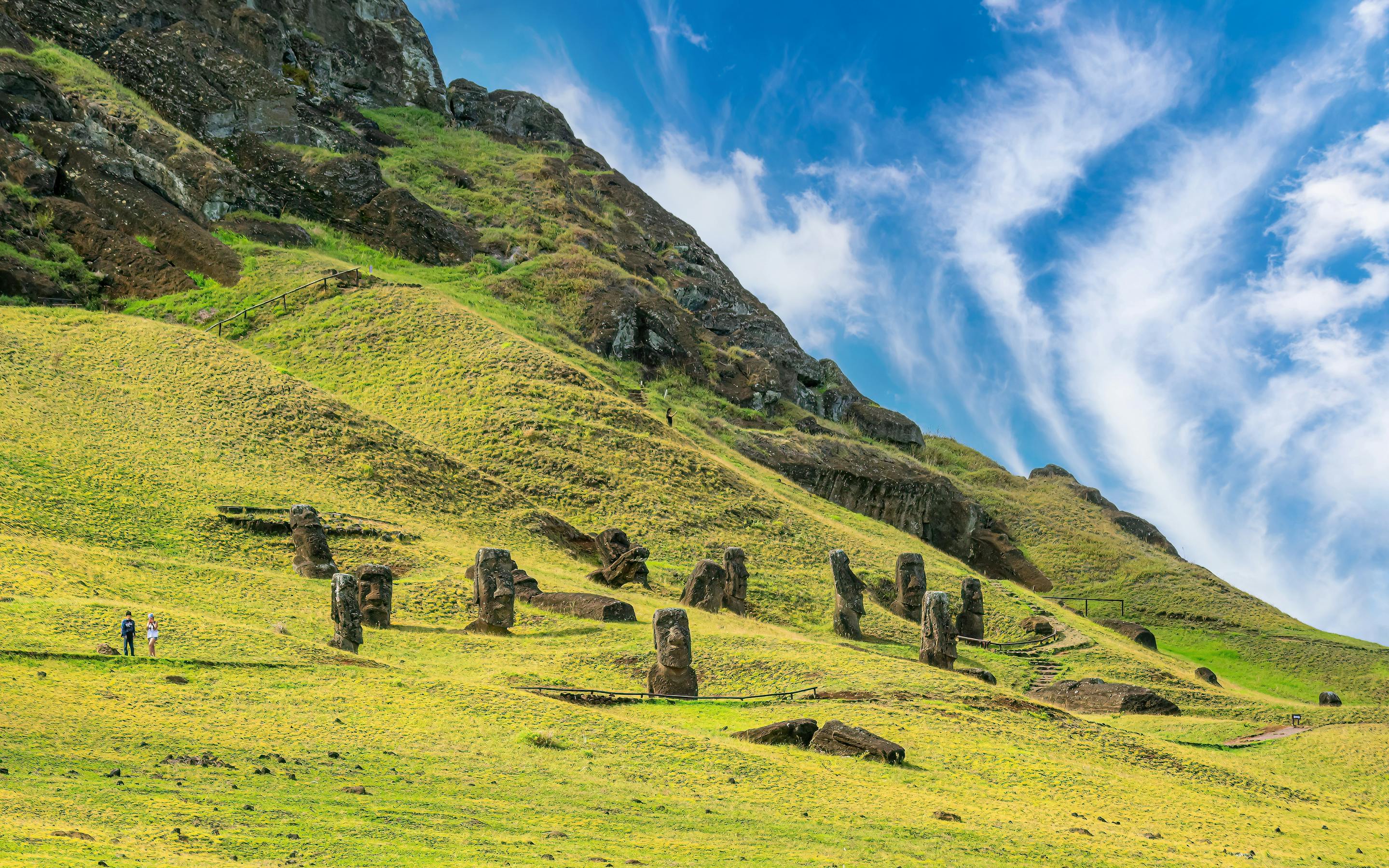 Stone moai statues lie scattered on a green hillside below a rocky crater wall at Rano Raraku on Easter Island.