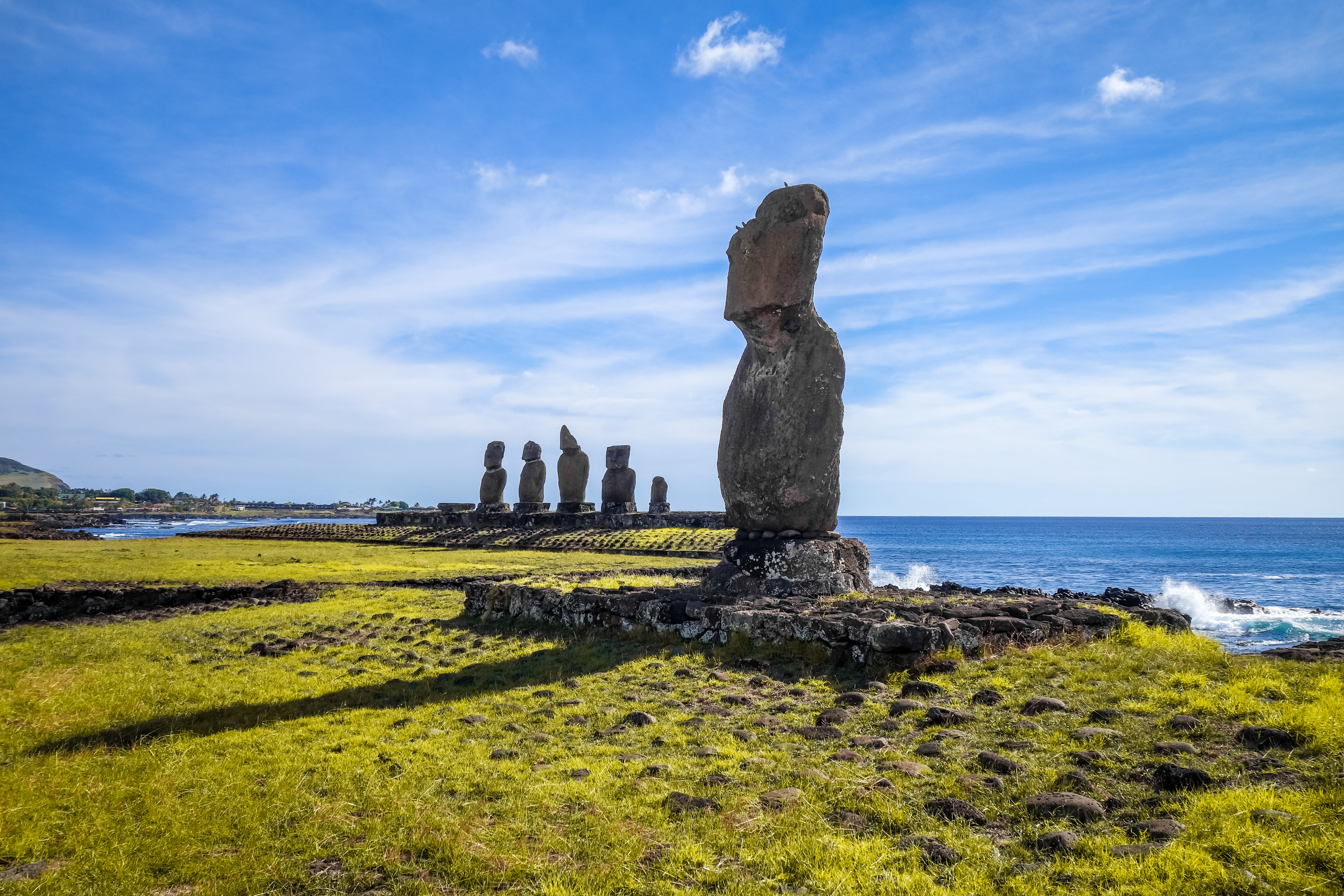 A lone moai stands near the rocky coast, with the Ahu Tongariki platform and ocean waves under a bright sky.