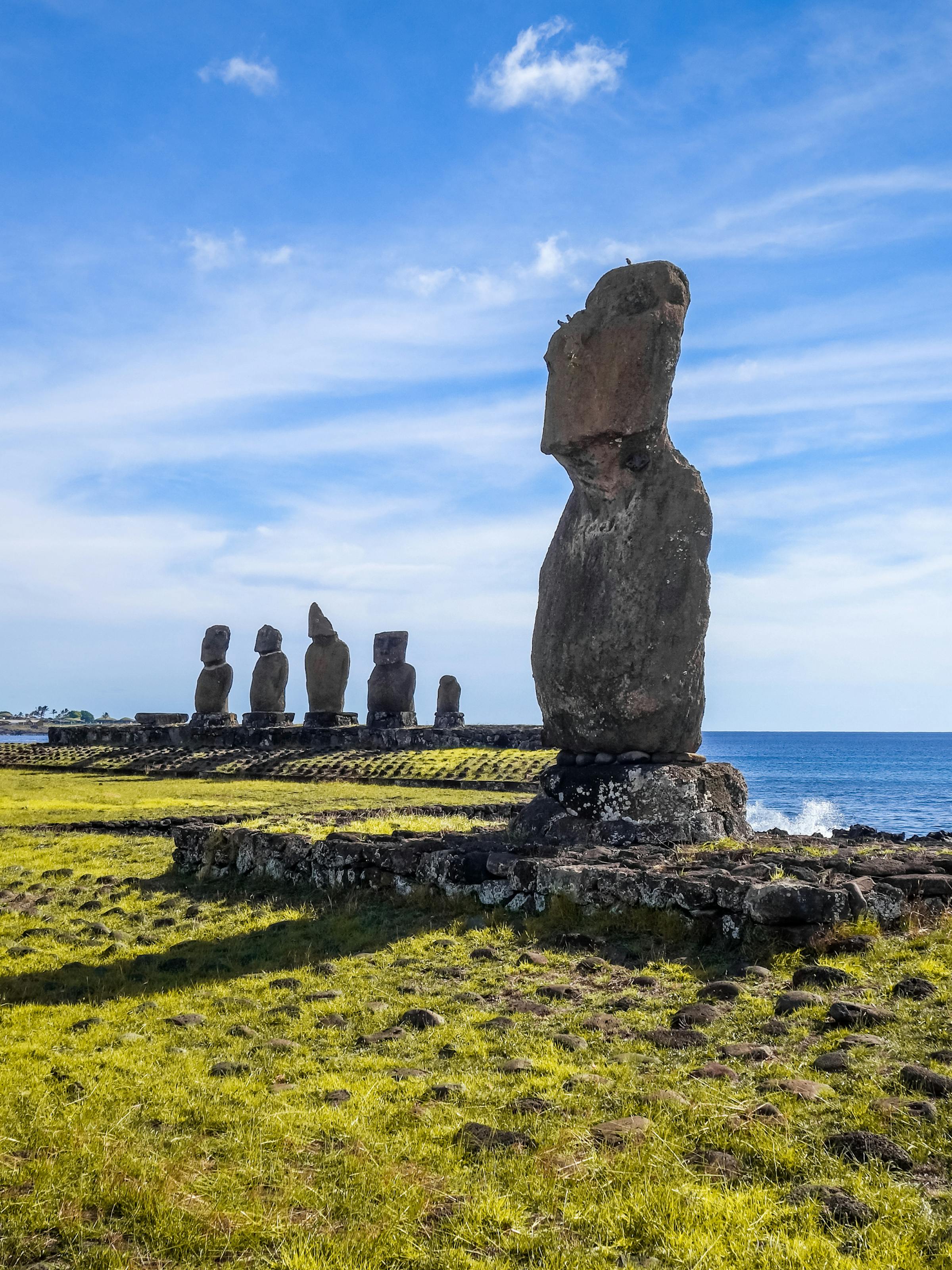 A lone moai stands near the rocky coast, with the Ahu Tongariki platform and ocean waves under a bright sky.