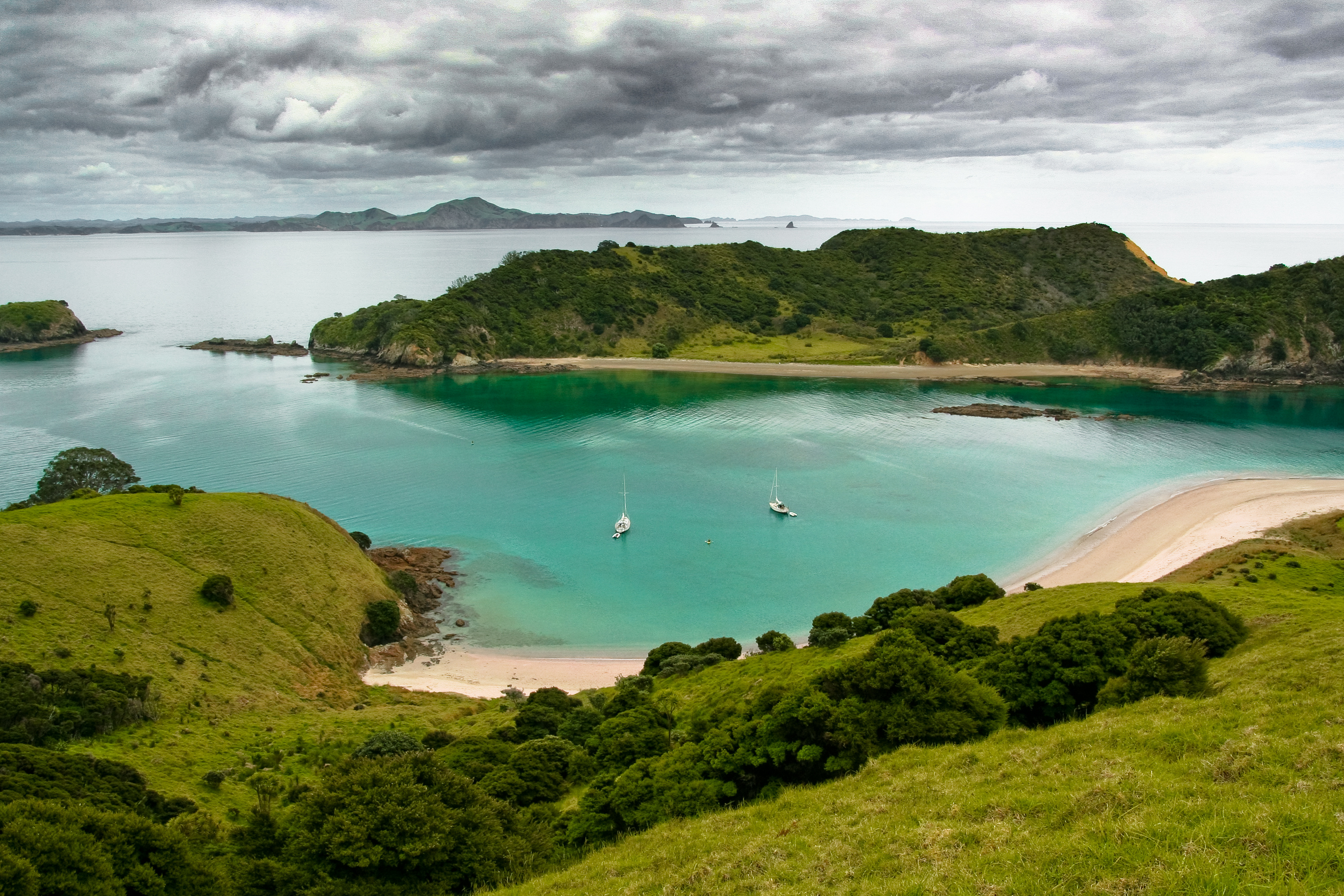 Green hills overlook a turquoise cove and pale beach in the Bay of Islands, with small sailboats anchored below.
