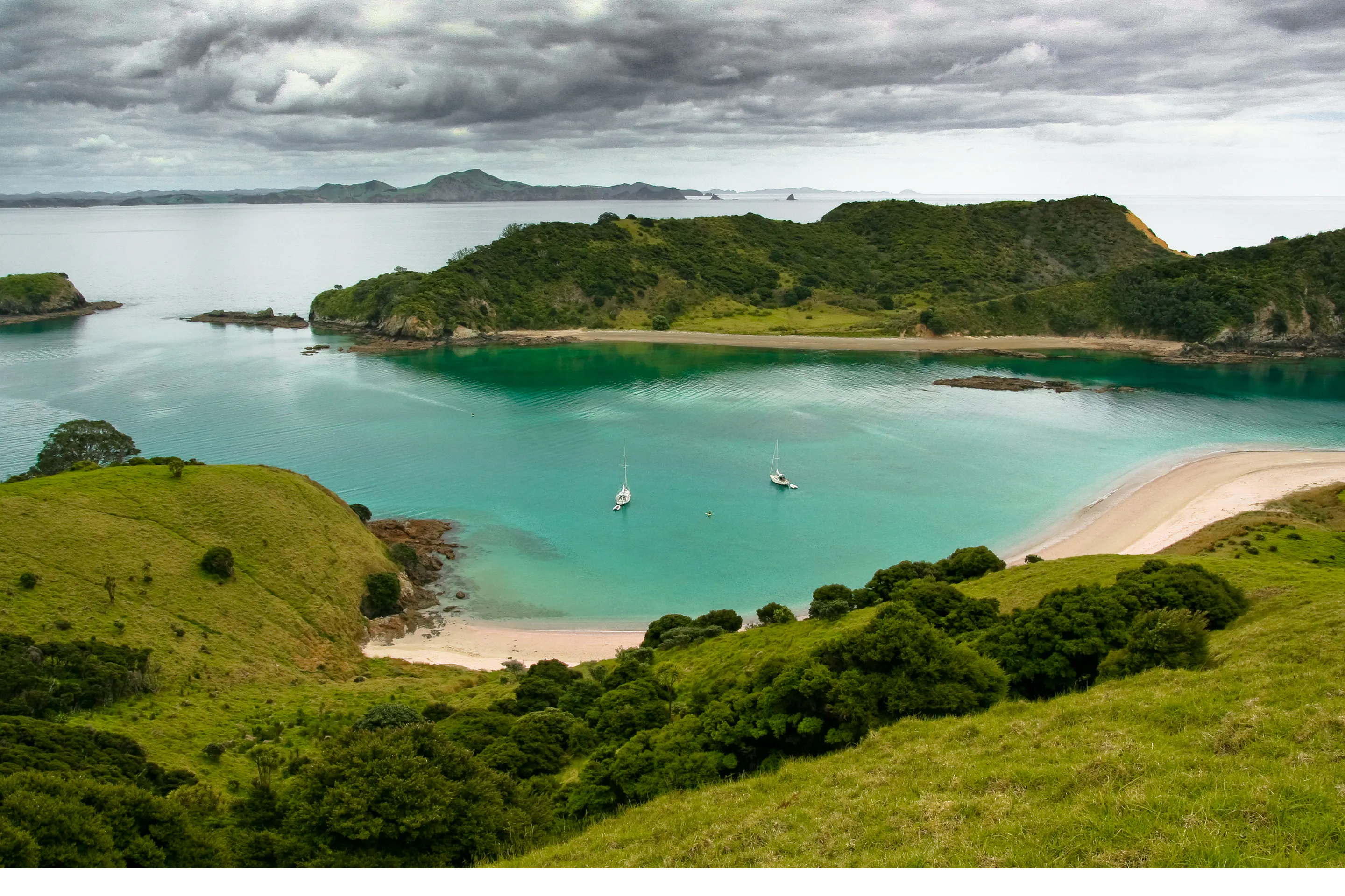 Green hills overlook a turquoise cove and pale beach in the Bay of Islands, with small sailboats anchored below.