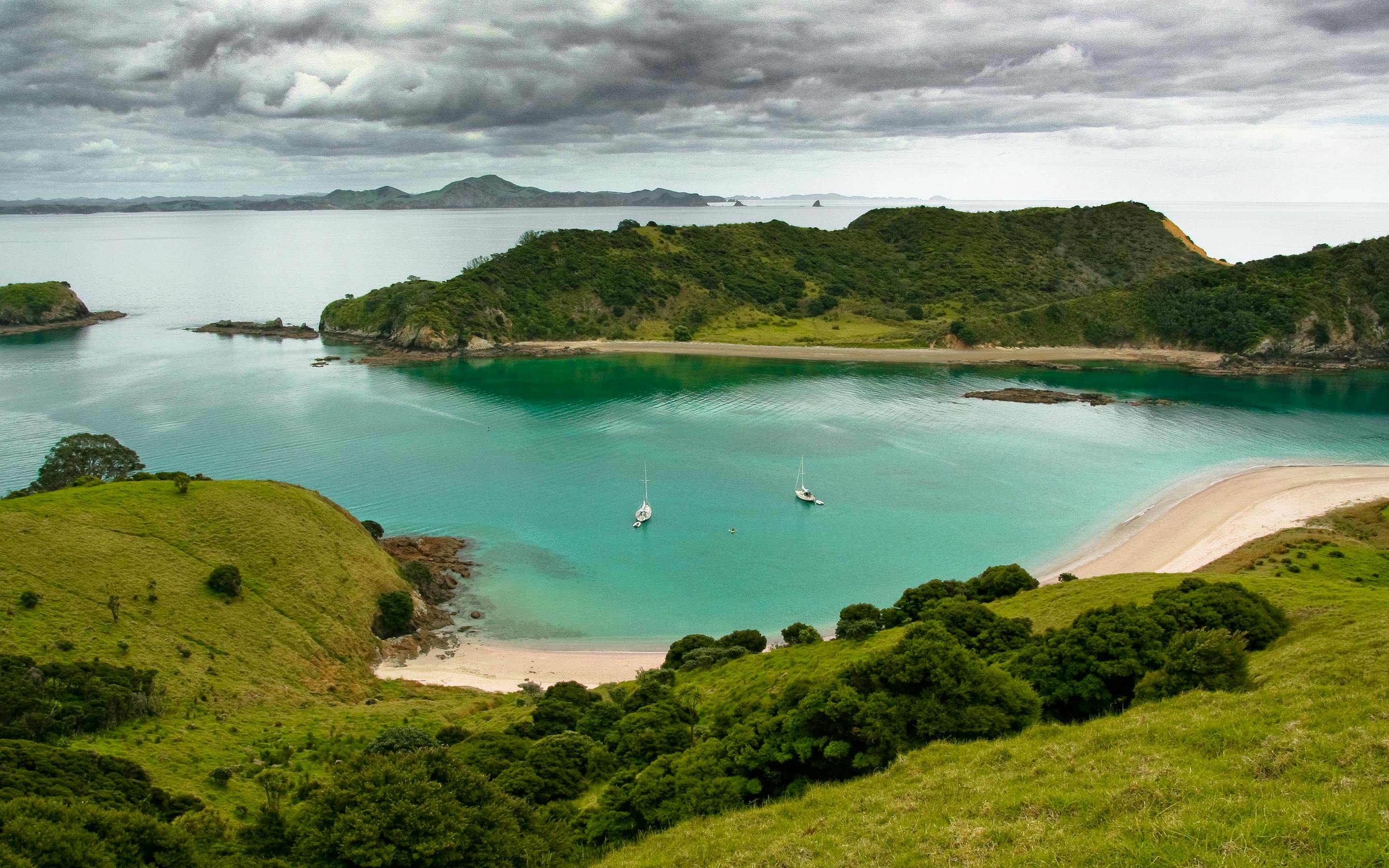 Green hills overlook a turquoise cove and pale beach in the Bay of Islands, with small sailboats anchored below.