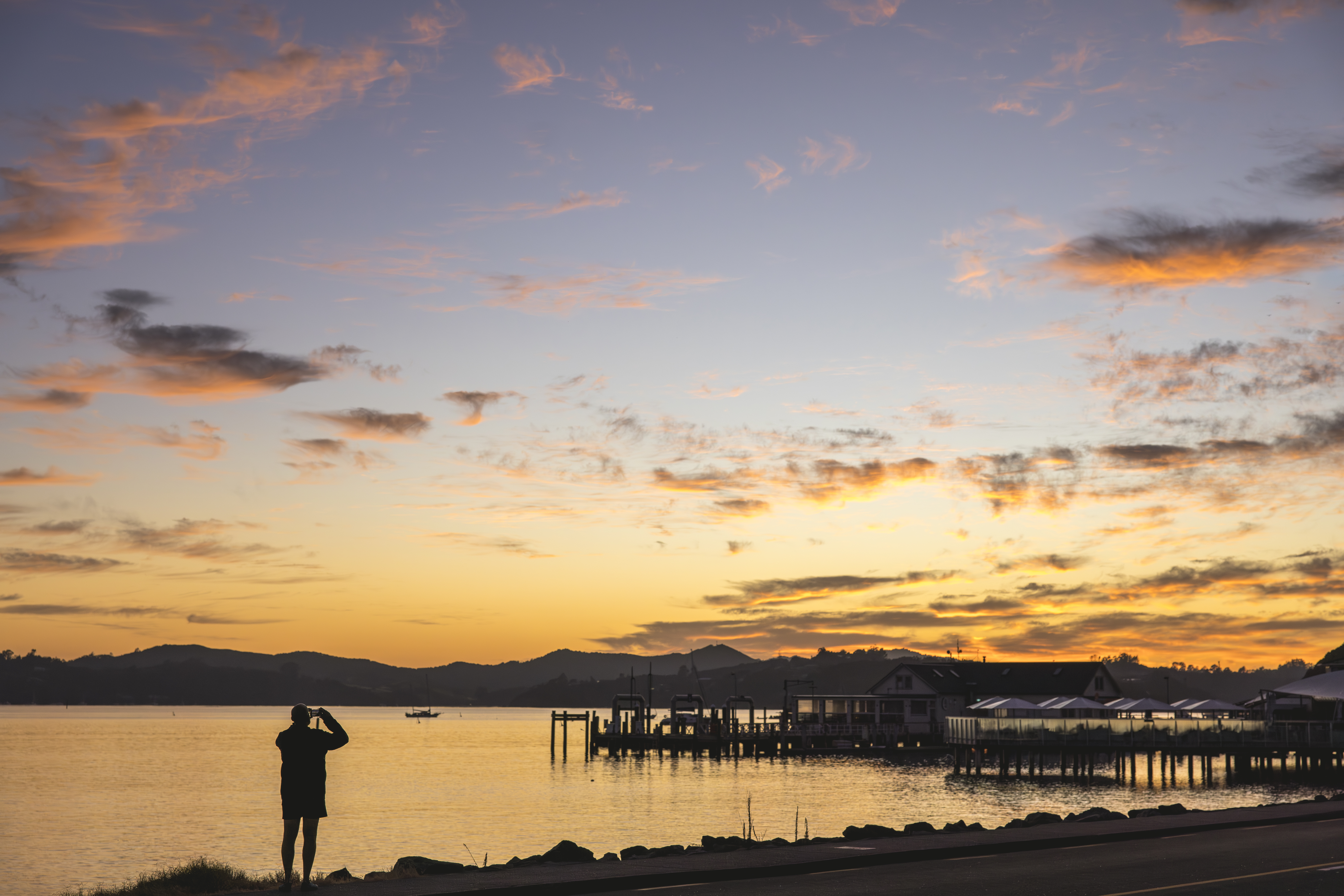 A person photographs a glowing sunset over calm water near a pier and waterfront buildings in Paihia town.