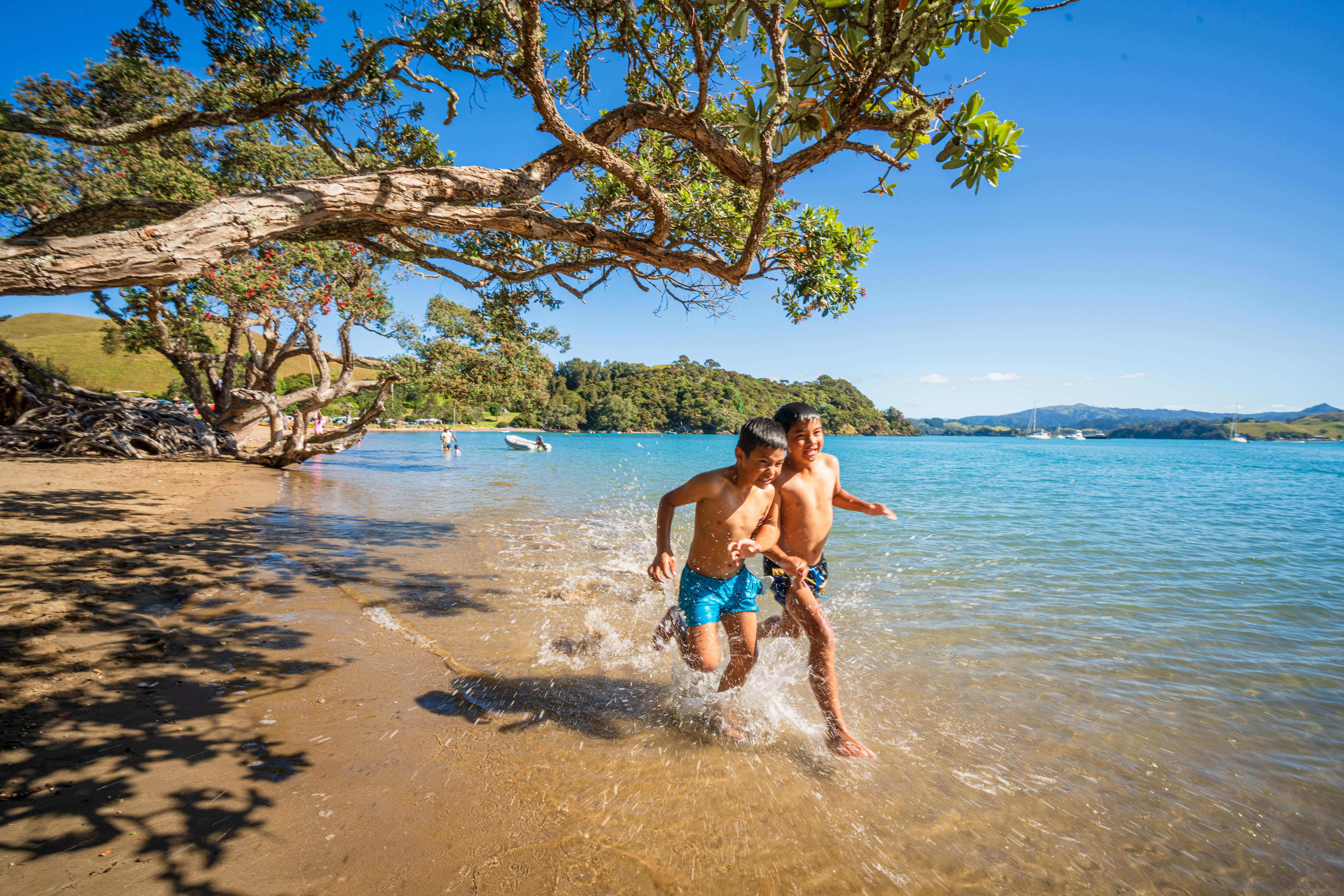 Two children run and splash along a shaded sandy beach in clear shallows, with calm water and hills beyond.