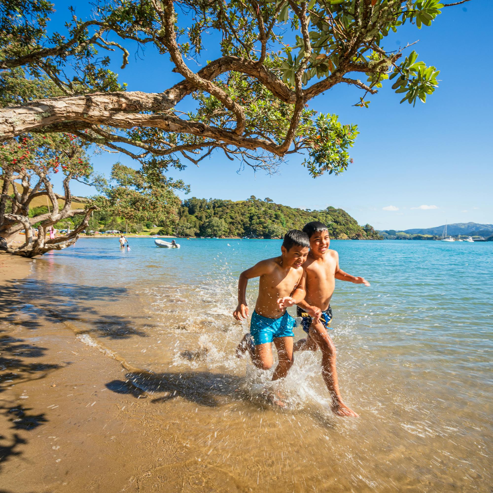Two children run and splash along a shaded sandy beach in clear shallows, with calm water and hills beyond.