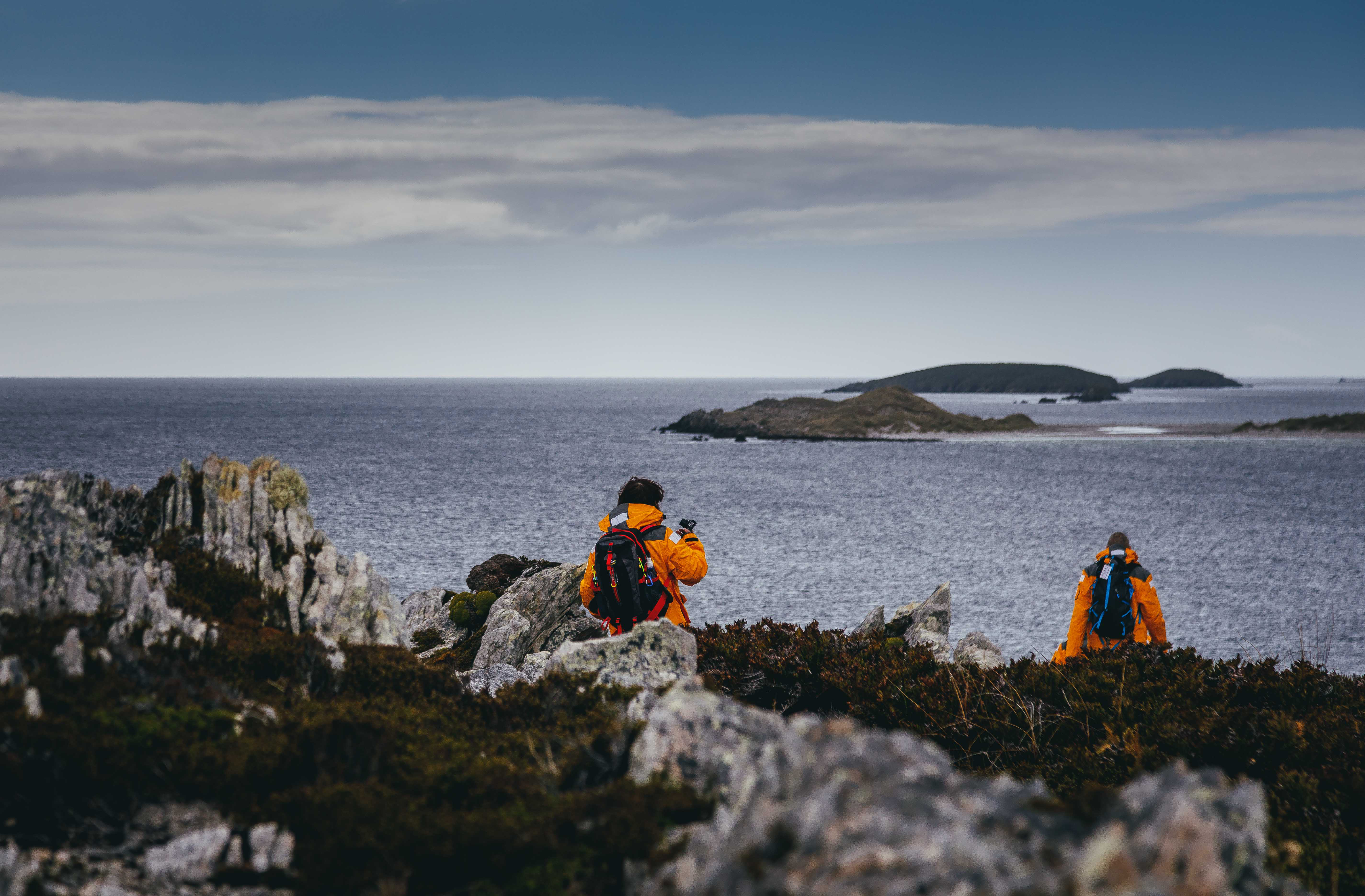 Two hikers in bright jackets walk among rocky coastal scrub, pausing to photograph the sea and islands beyond.