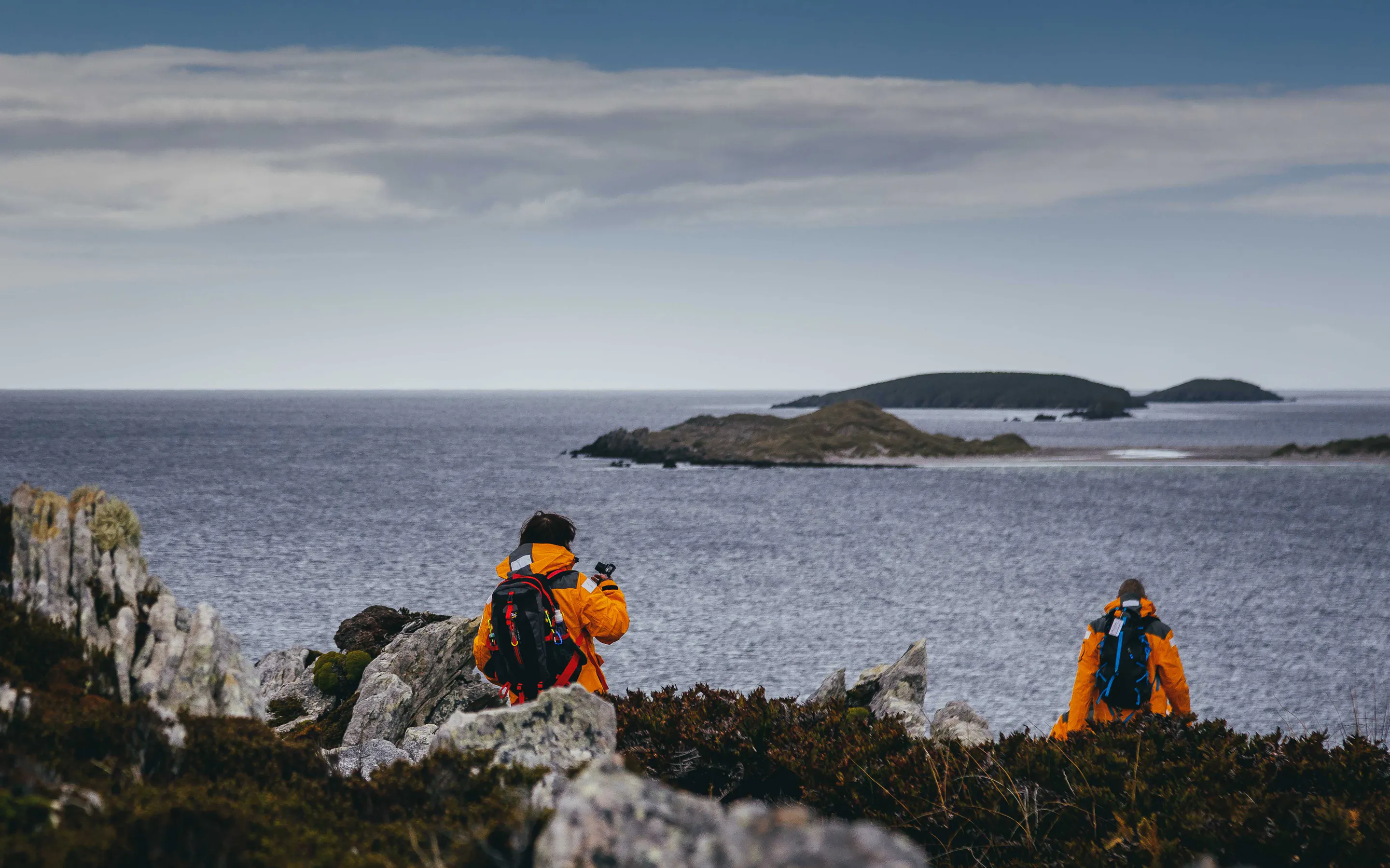 Two hikers in bright jackets walk among rocky coastal scrub, pausing to photograph the sea and islands beyond.