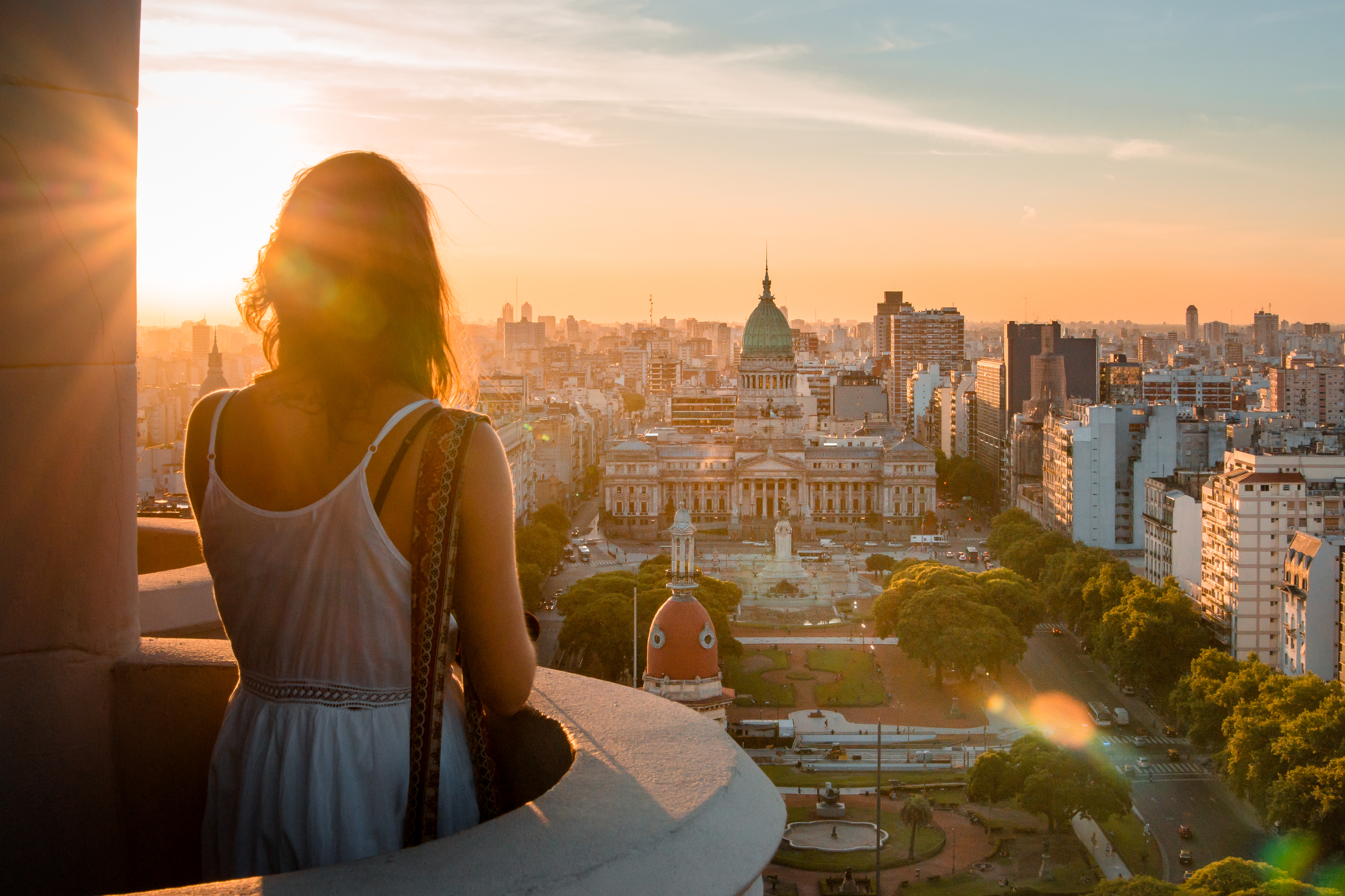 A woman stands on a balcony at sunset, looking over Buenos Aires toward the domed Congress building and city streets.