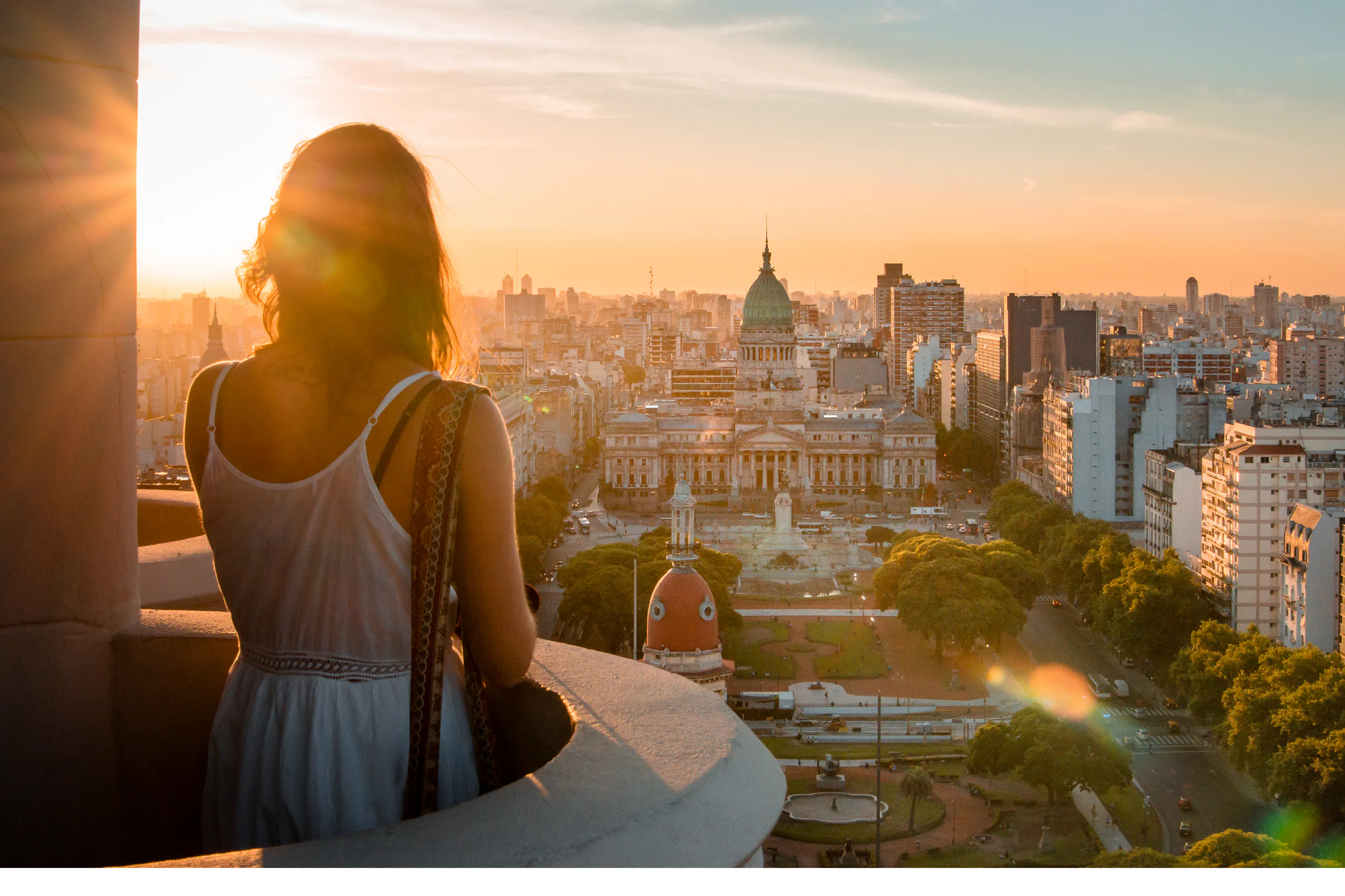 A woman stands on a balcony at sunset, looking over Buenos Aires toward the domed Congress building and city streets.