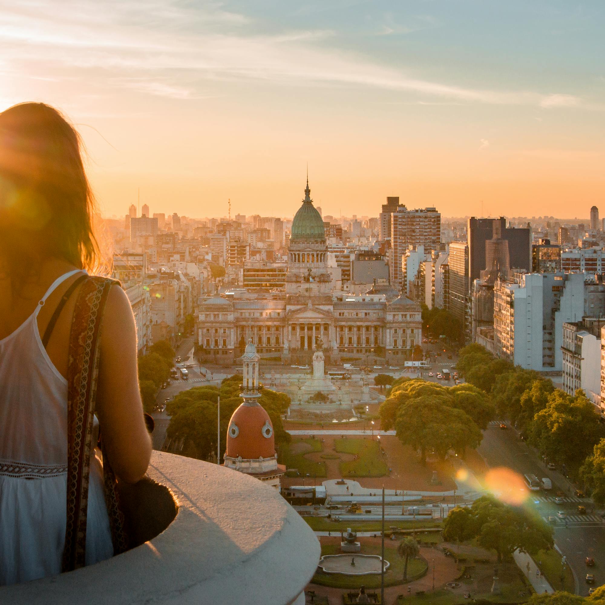 A woman stands on a balcony at sunset, looking over Buenos Aires toward the domed Congress building and city streets.
