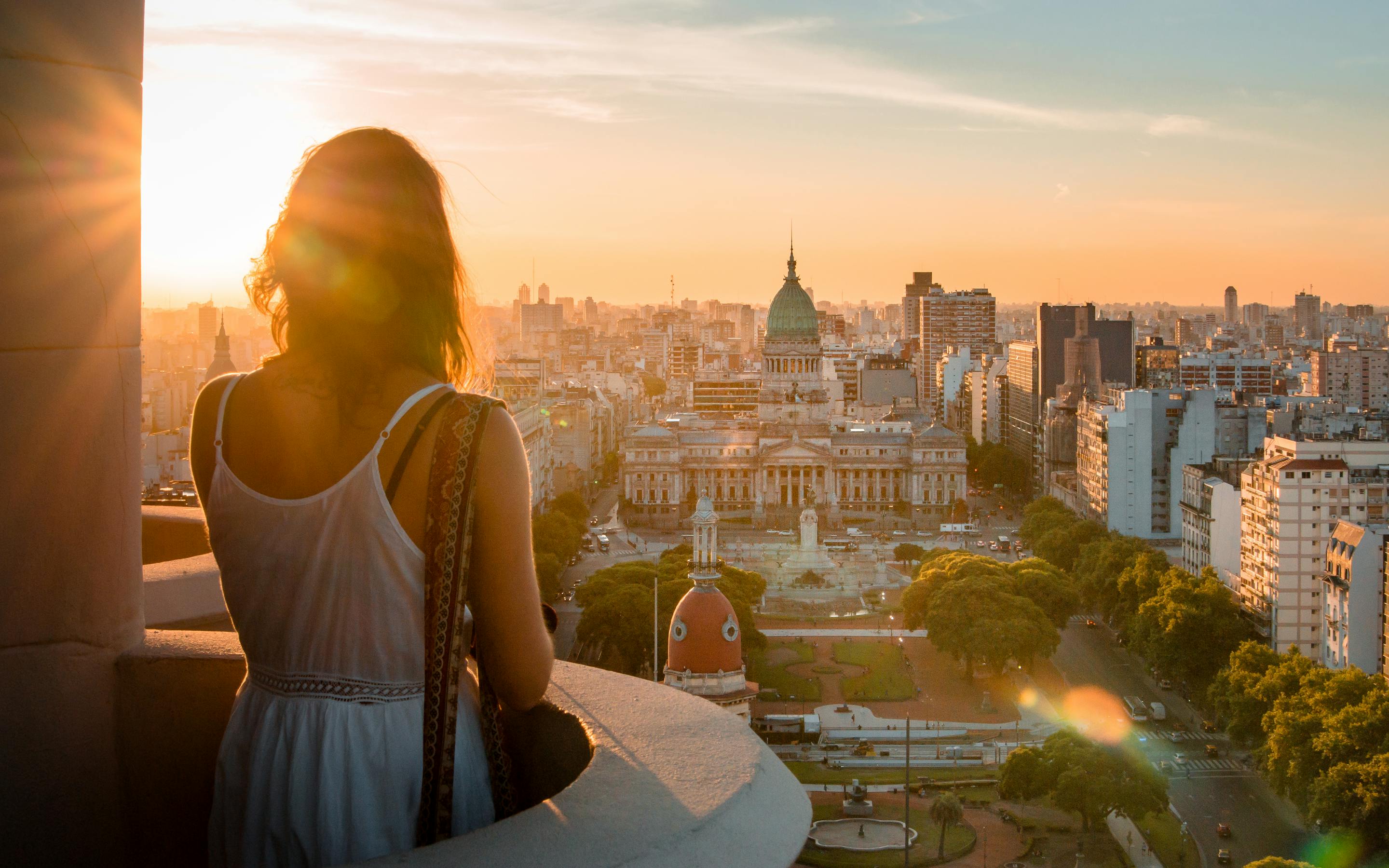 A woman stands on a balcony at sunset, looking over Buenos Aires toward the domed Congress building and city streets.