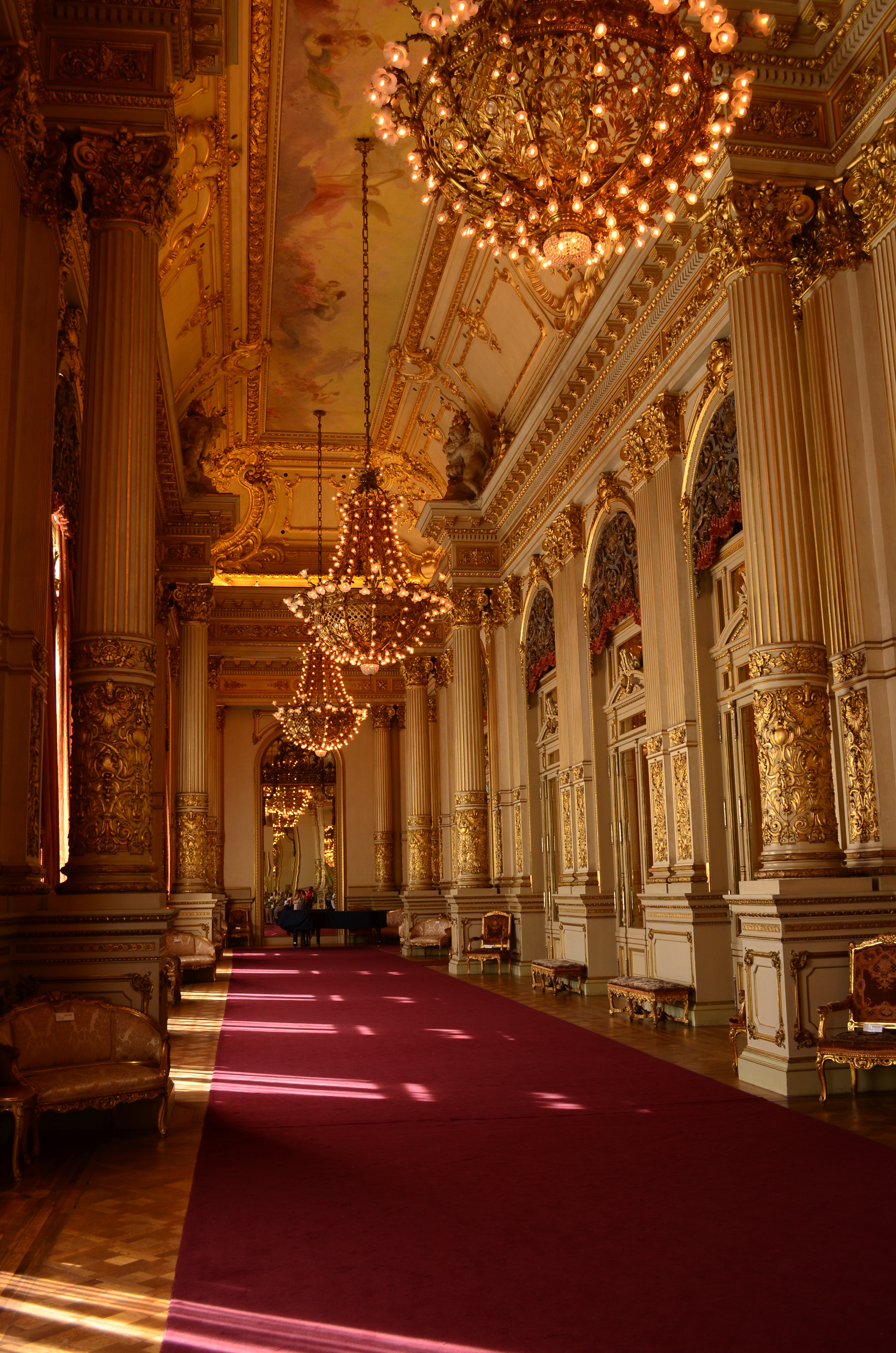 Golden chandeliers hang above a red-carpeted corridor, with ornate columns and mirrors in a grand Buenos Aires interior.