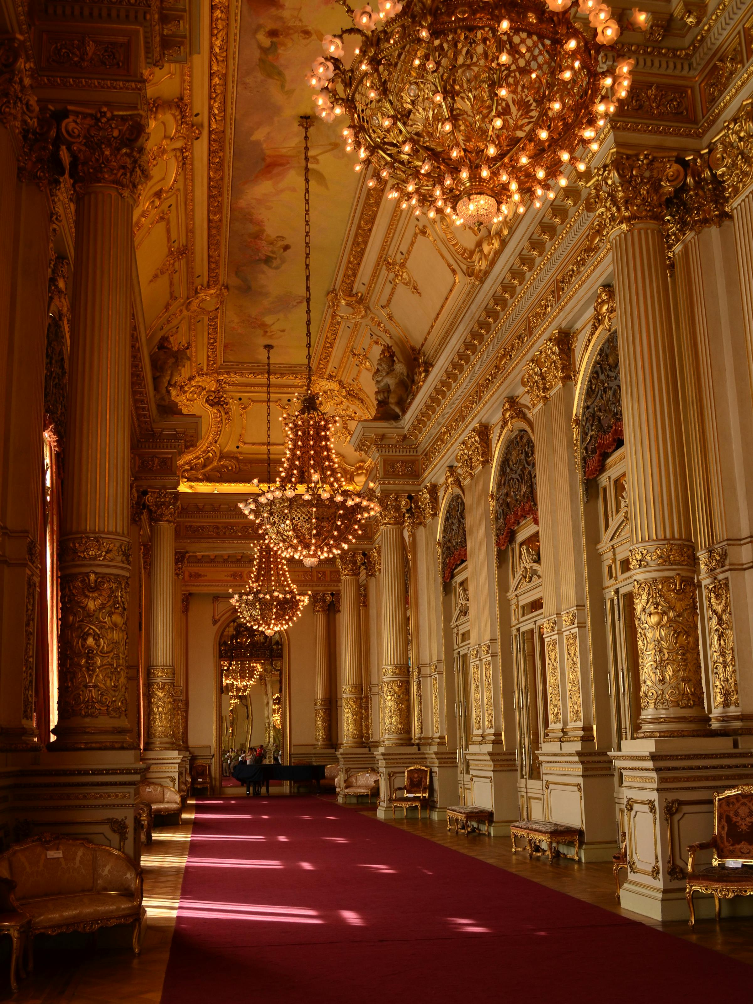 Golden chandeliers hang above a red-carpeted corridor, with ornate columns and mirrors in a grand Buenos Aires interior.