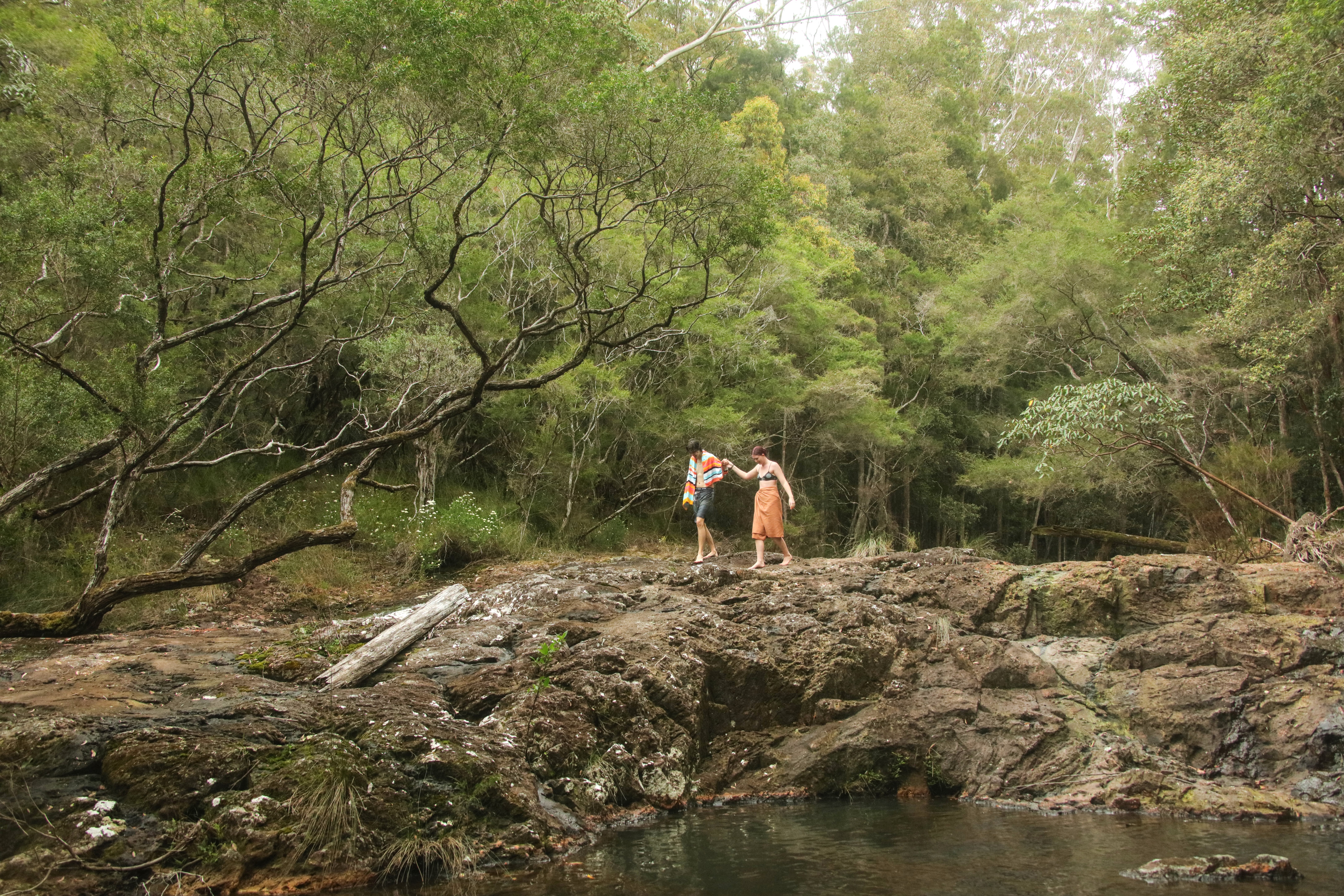 Two hikers stand on a rocky creek bed beneath open woodland, surrounded by green trees and dappled shade.