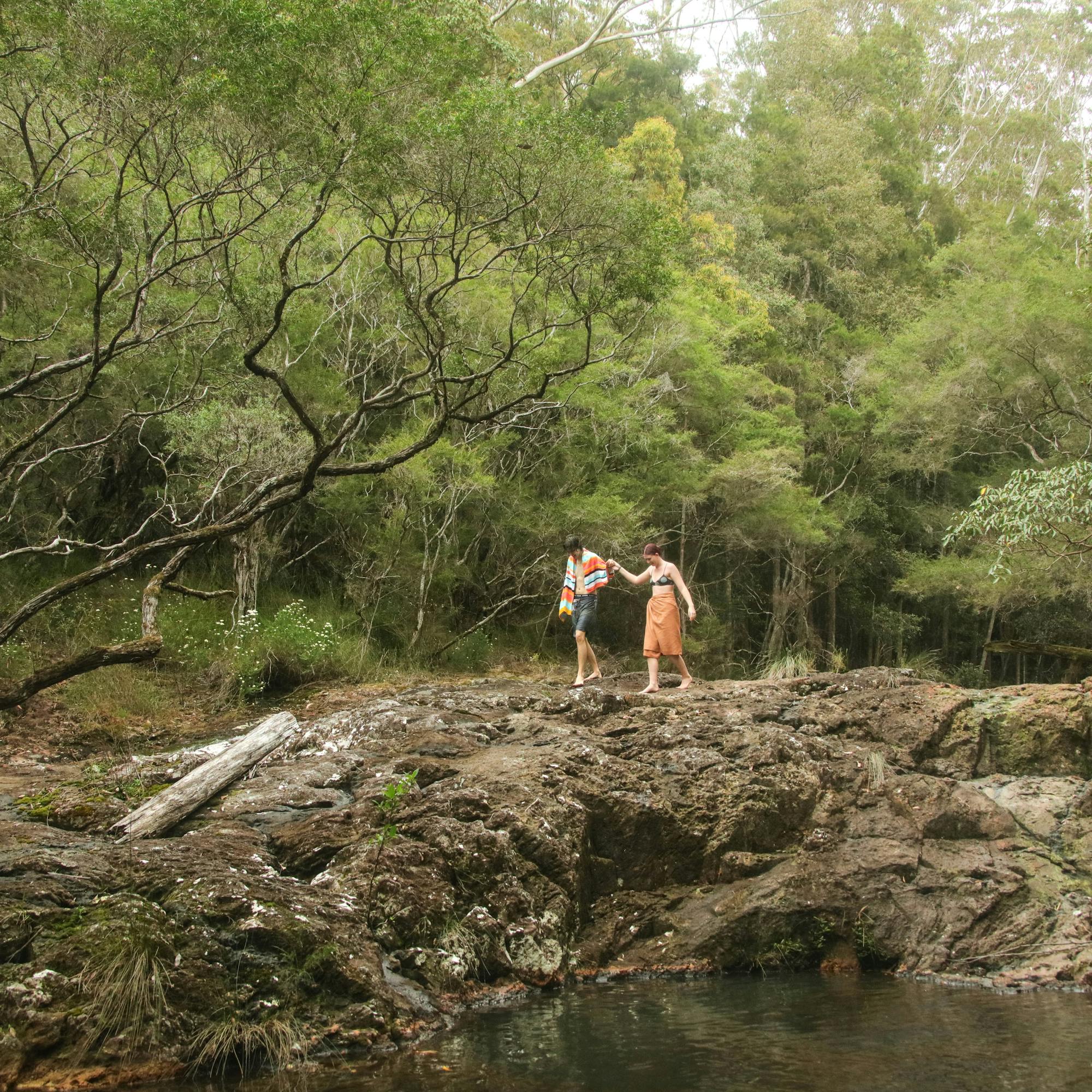 Two hikers stand on a rocky creek bed beneath open woodland, surrounded by green trees and dappled shade.