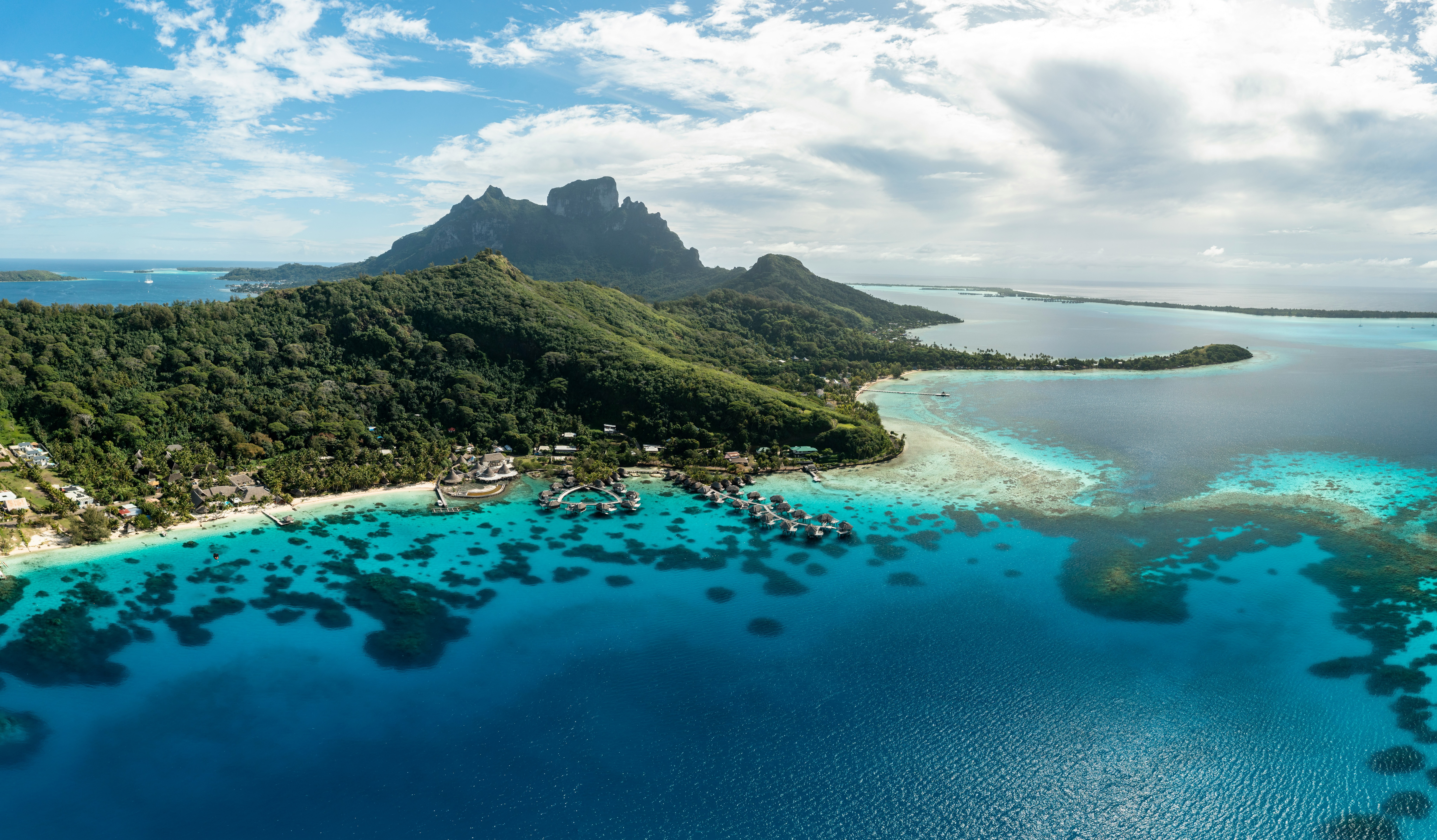 An aerial view shows a lush island ringed by turquoise lagoon and coral reef, with sandbars and deep blue ocean beyond.