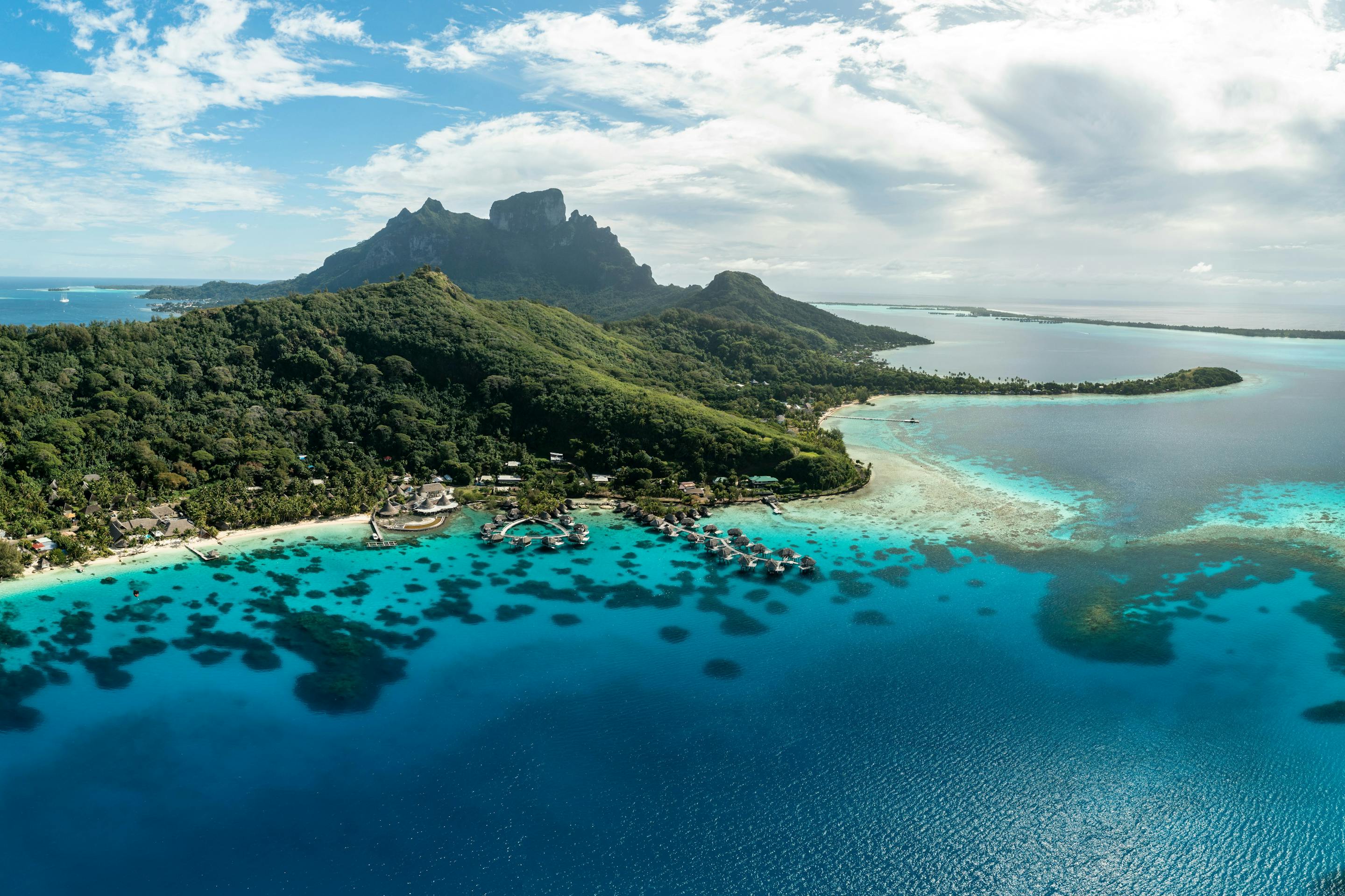 An aerial view shows a lush island ringed by turquoise lagoon and coral reef, with sandbars and deep blue ocean beyond.