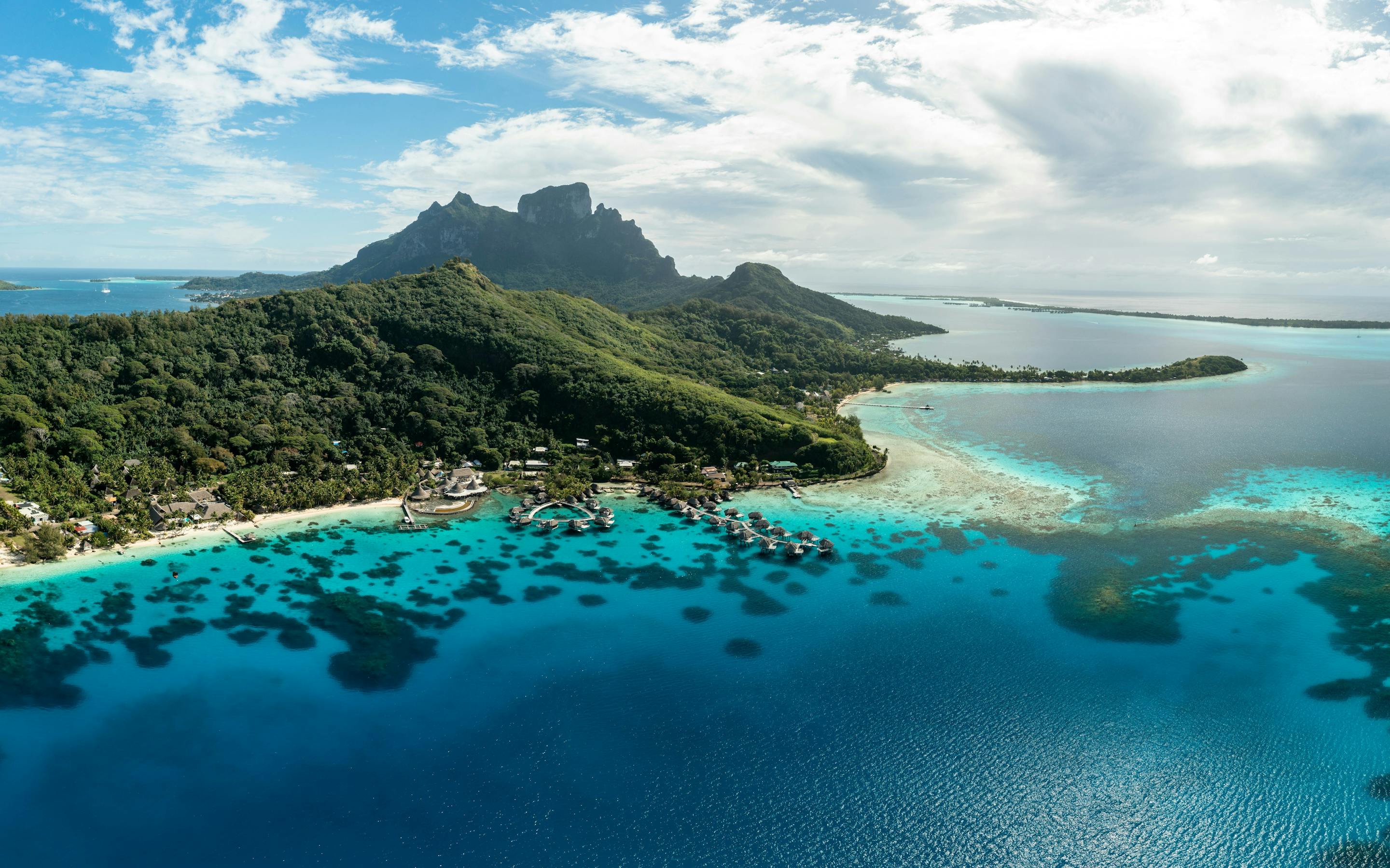 An aerial view shows a lush island ringed by turquoise lagoon and coral reef, with sandbars and deep blue ocean beyond.