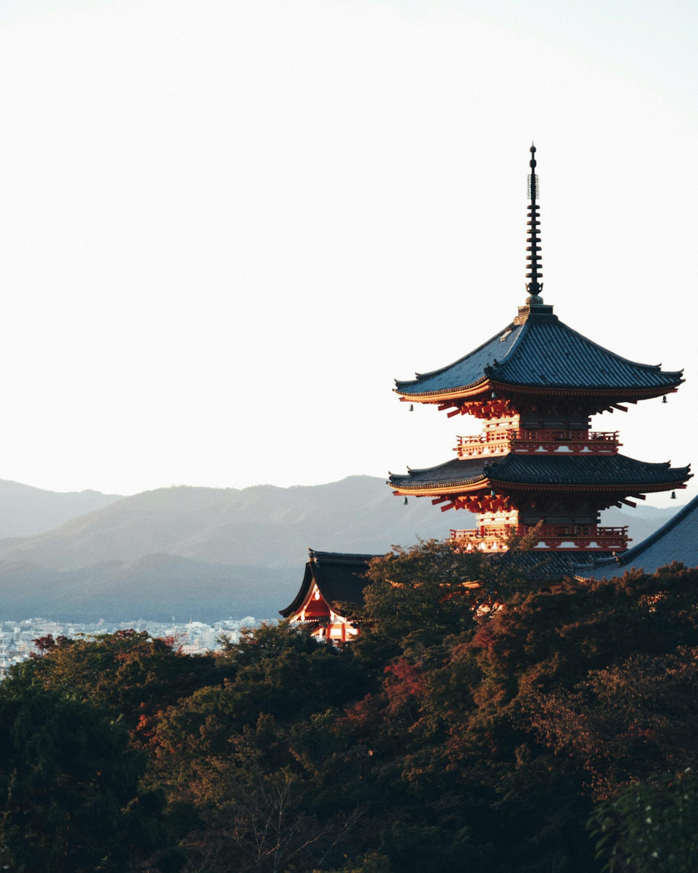 Multi-tiered Japanese pagoda rising above autumn-colored trees, with mountains and a city visible in the distance at sunset.