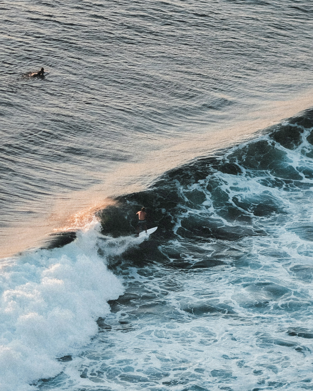 Aerial view of a surfer riding a breaking wave, with another surfer waiting farther out in the ocean.