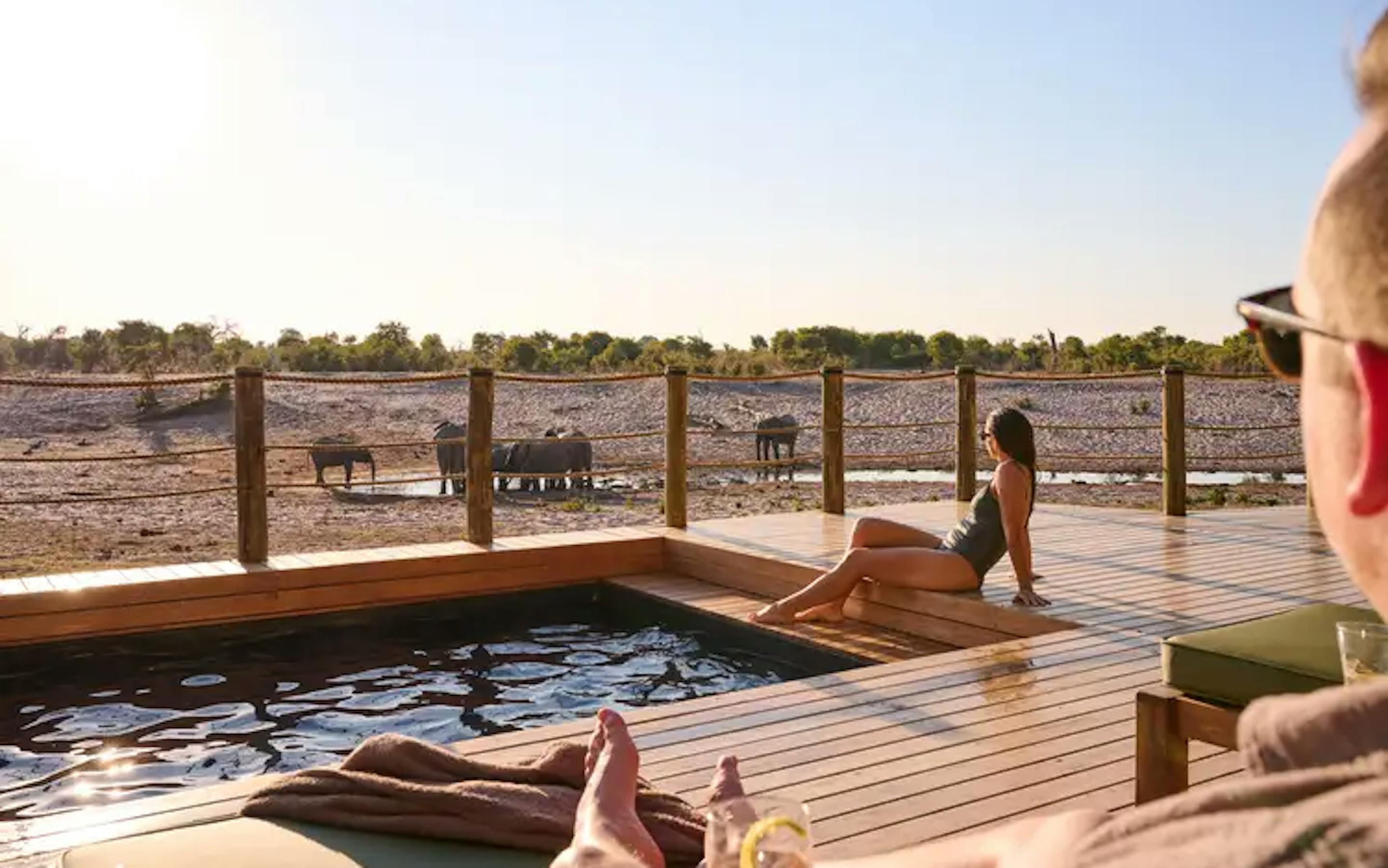 Two people in swimsuits lay by a pool, with wildlife in the distance.